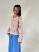 A woman wearing a taupe textured knit sweater with blue patterns and orange butterfly appliqués, paired with wide-leg blue pants, standing against a plain light gray studio background.