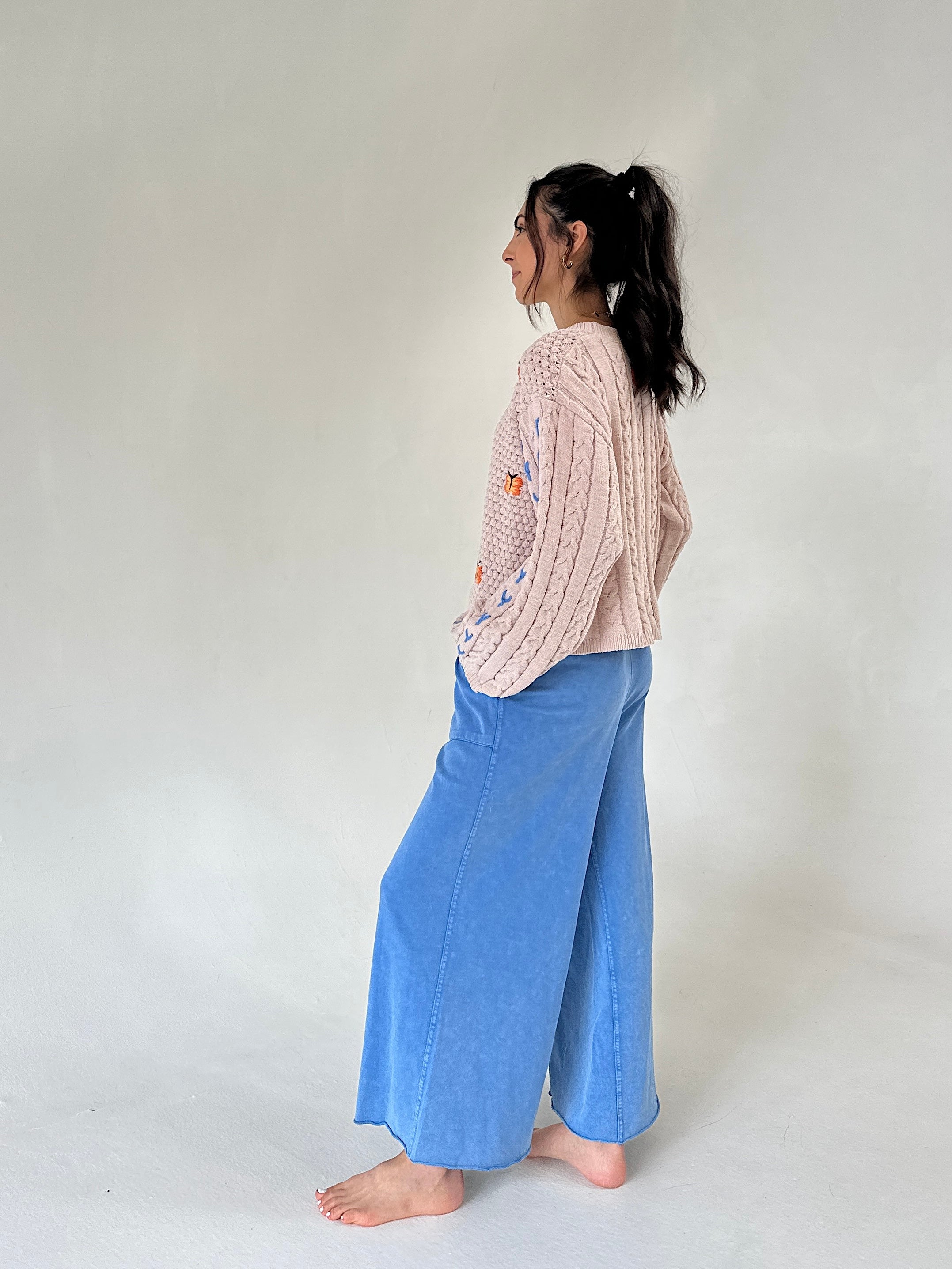 A woman stands in profile wearing a light taupe cable-knit sweater with colorful embroidery, paired with wide-leg blue pants, against a plain light gray studio background.