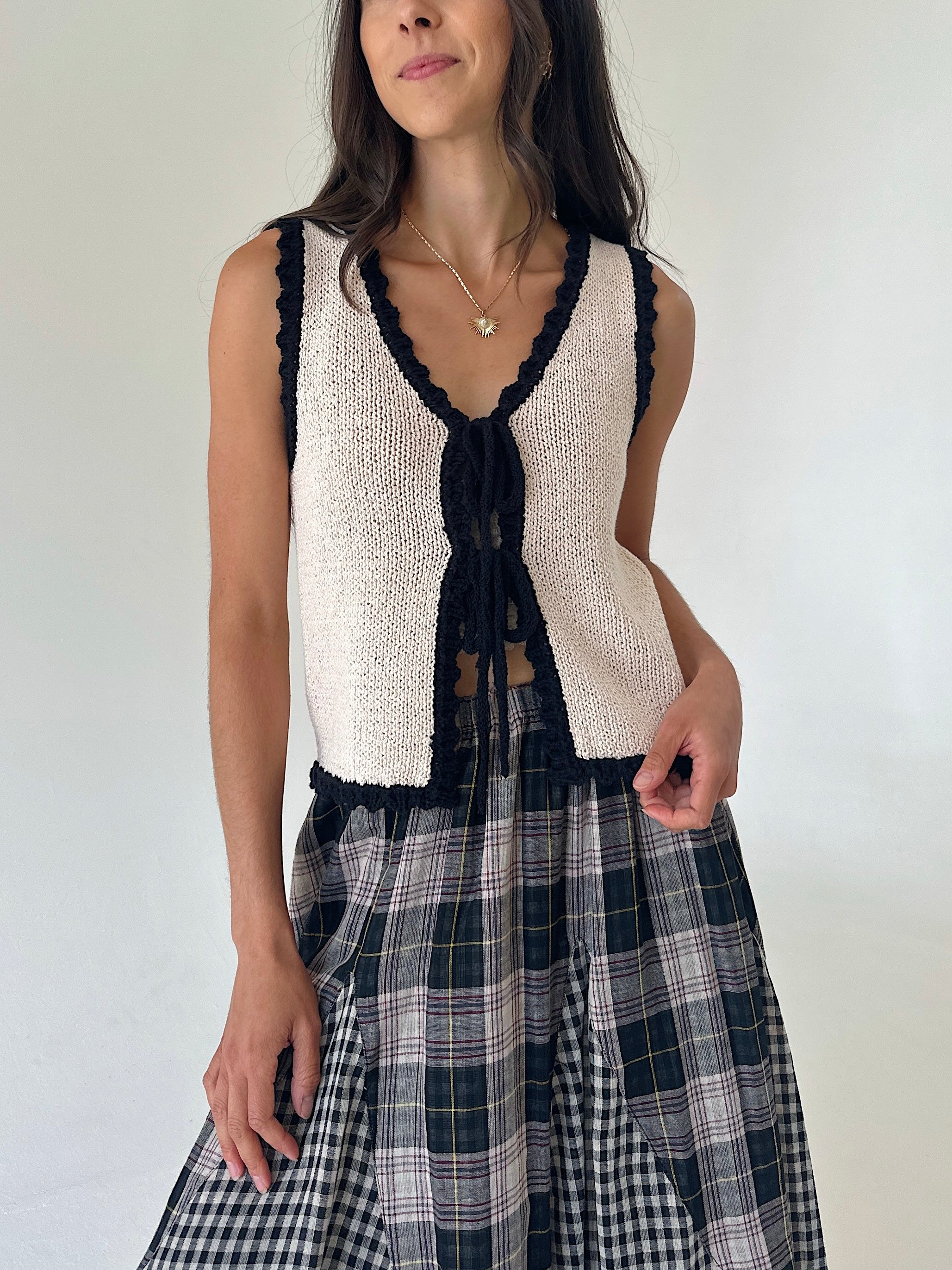 A woman wearing a sleeveless cream knit vest with black trim and a black front tie, paired with a black, white, and yellow plaid skirt, against a plain light background.