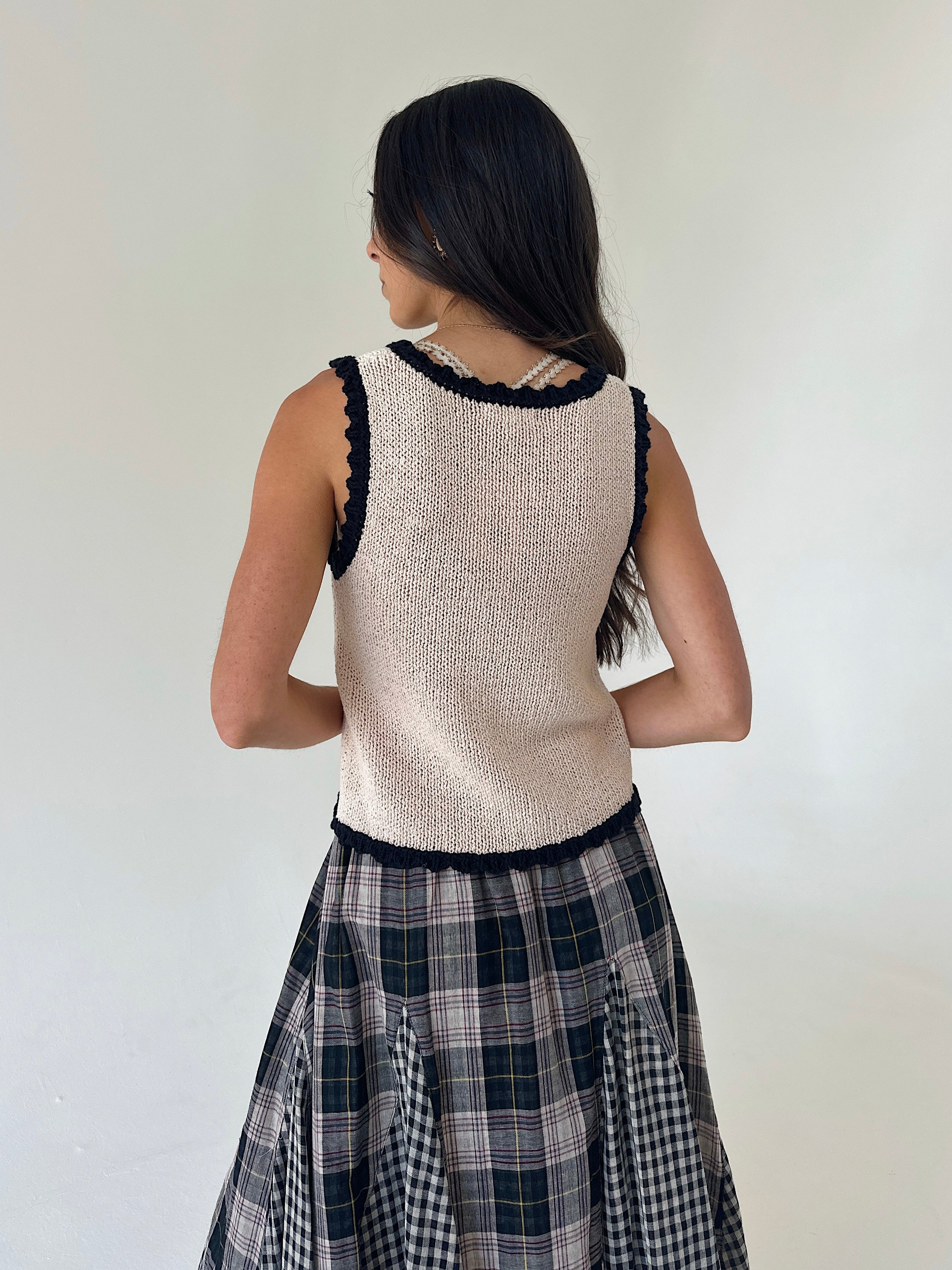 A woman shown from the back wearing a sleeveless cream knit vest with black lace trim and a black-and-white plaid skirt, against a plain off-white wall.
