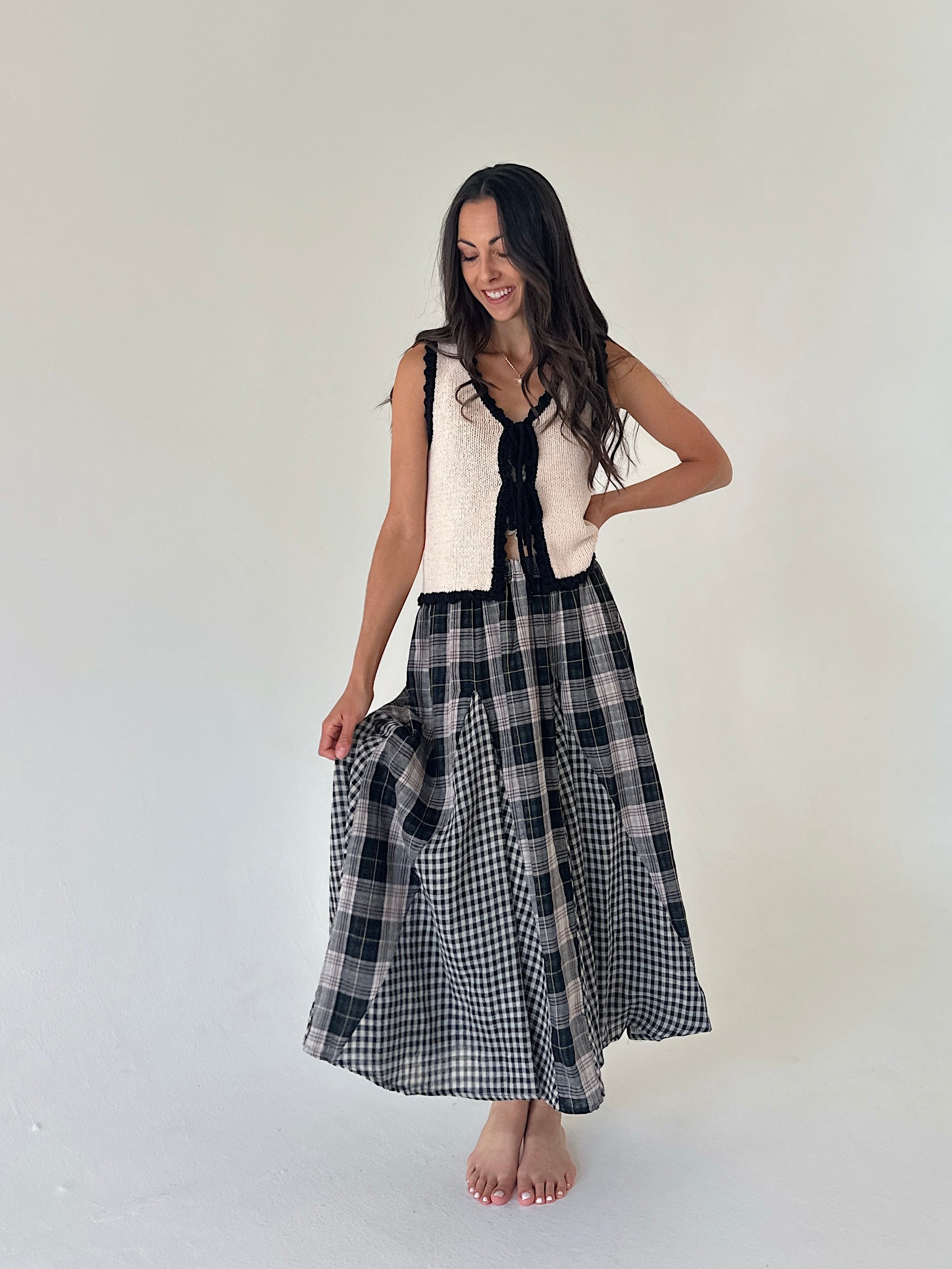 A woman modeling a sleeveless cream knit vest with a black ribbon tie and a black and gray plaid maxi skirt, standing against a plain off-white studio background.