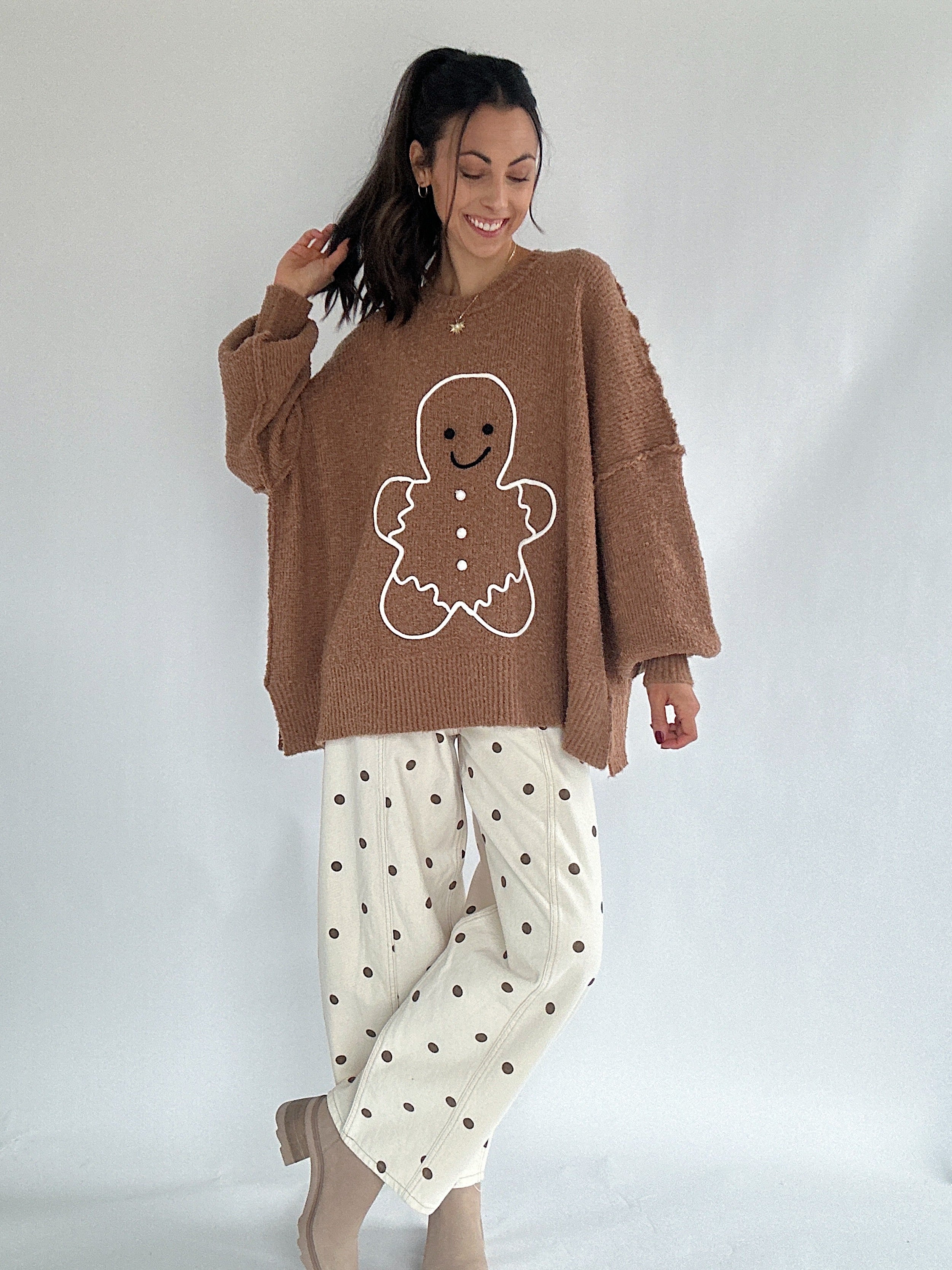 Woman wearing a brown sweater with a gingerbread man design and white pants with black dots on a plain background