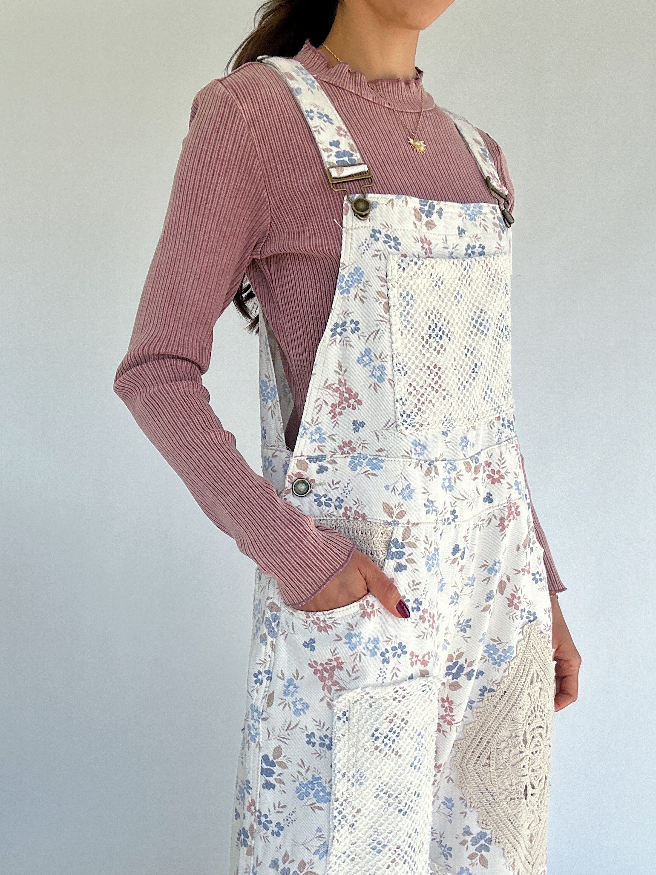 Person wearing floral overalls over a pink ribbed sweater against a plain background showing side pockets