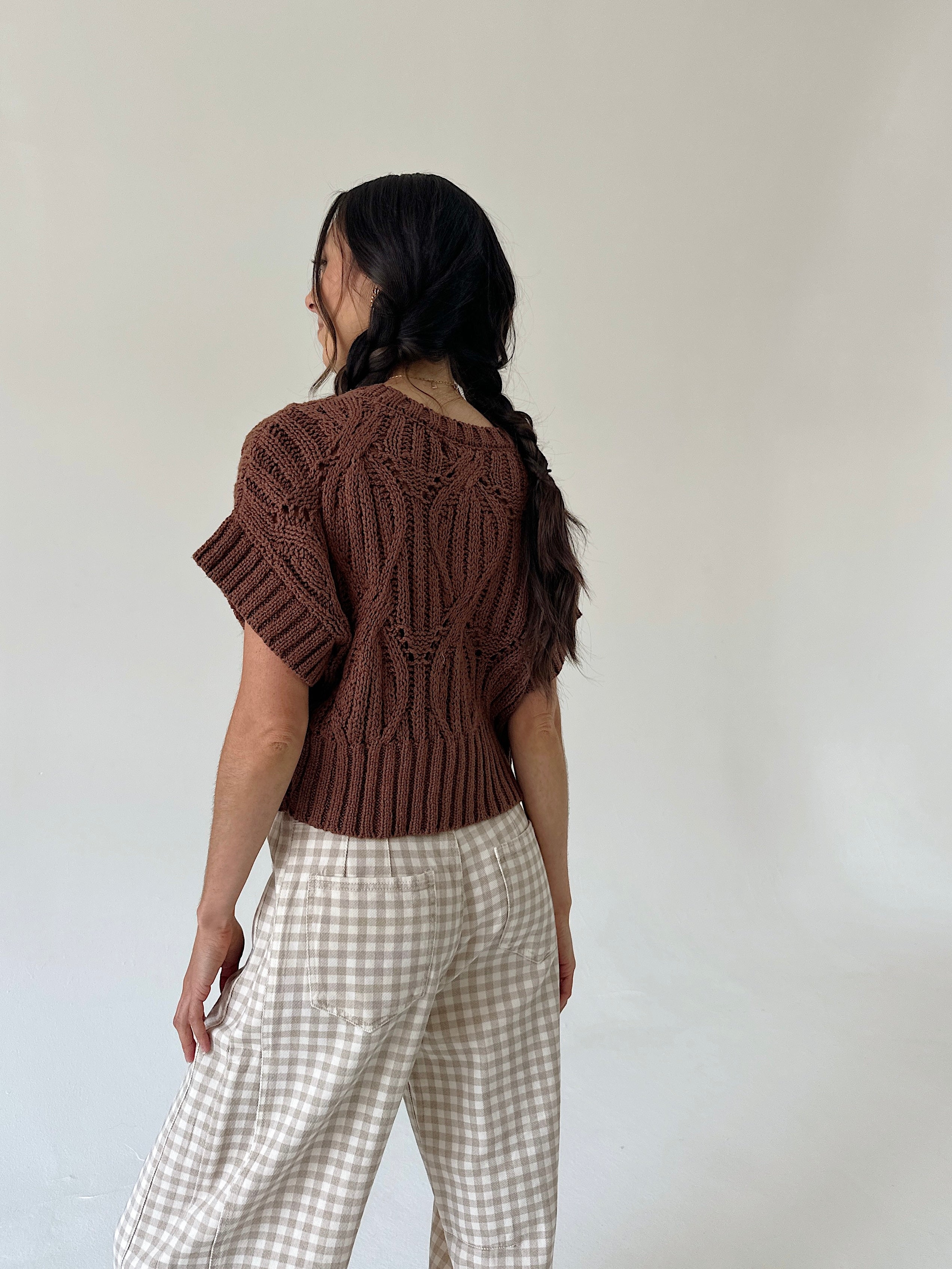 A woman shown from the back wearing a brown short-sleeve knit top and beige-and-white gingham pants against a plain off-white wall.