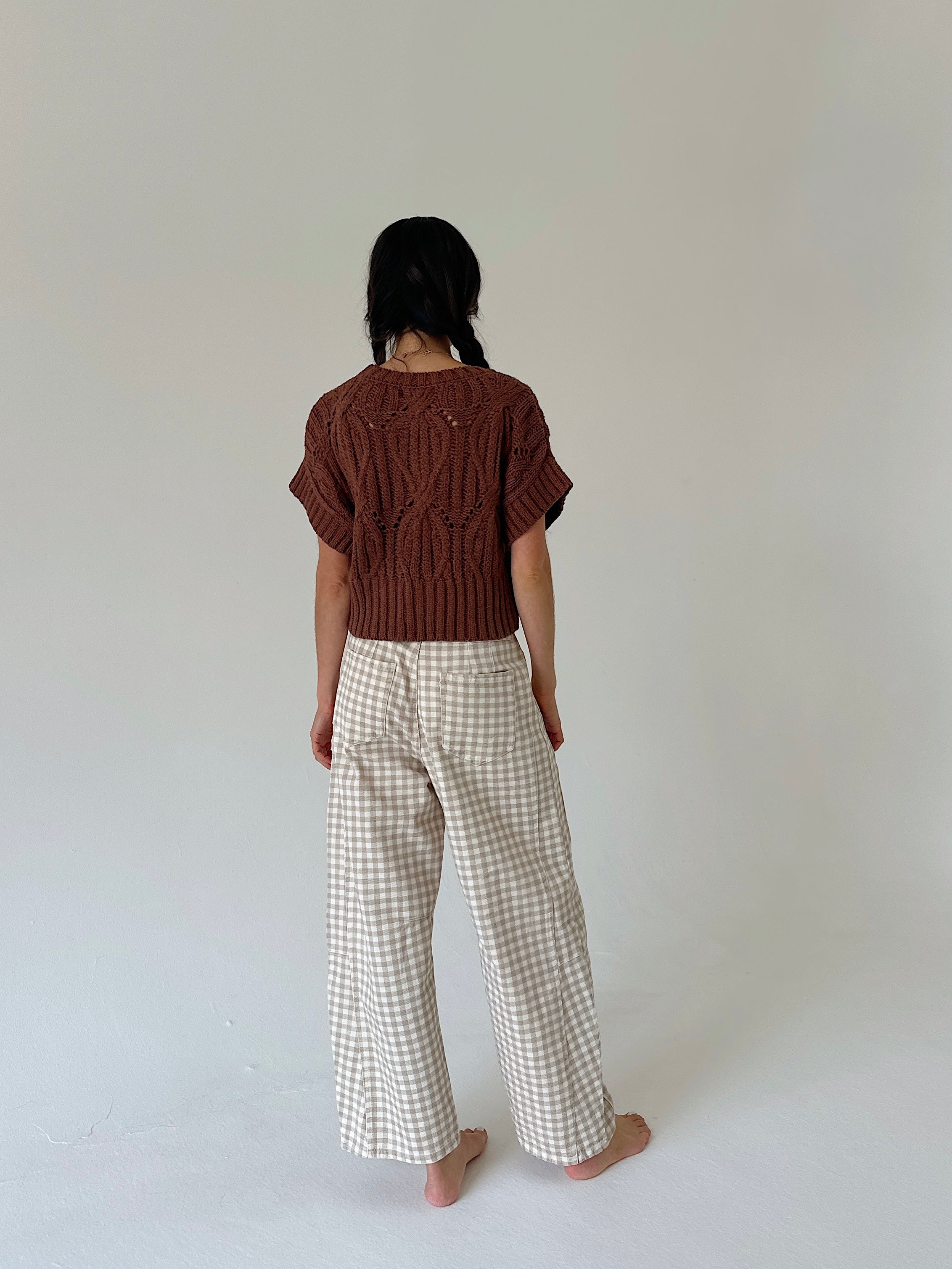 A person standing with their back to the camera, wearing a brown short-sleeve knit top and beige-and-white gingham wide-leg trousers, against a plain light gray studio background.
