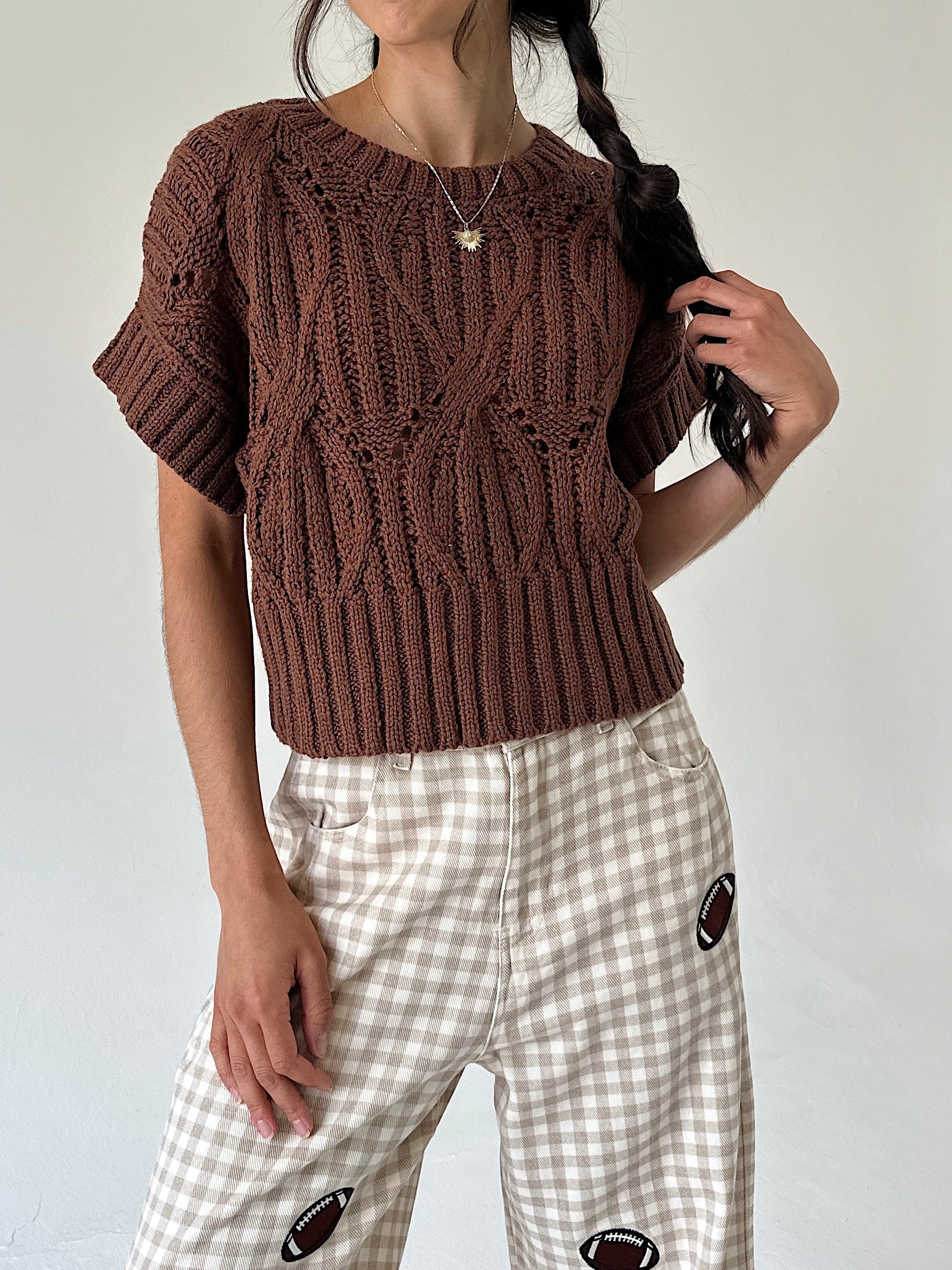 A person wearing a brown short-sleeve cable-knit sweater and beige-and-white gingham pants with football patch details, photographed against a plain light background.