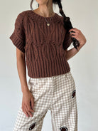 A person wearing a brown short-sleeve cable-knit sweater and beige-and-white gingham pants with football patch details, photographed against a plain light background.