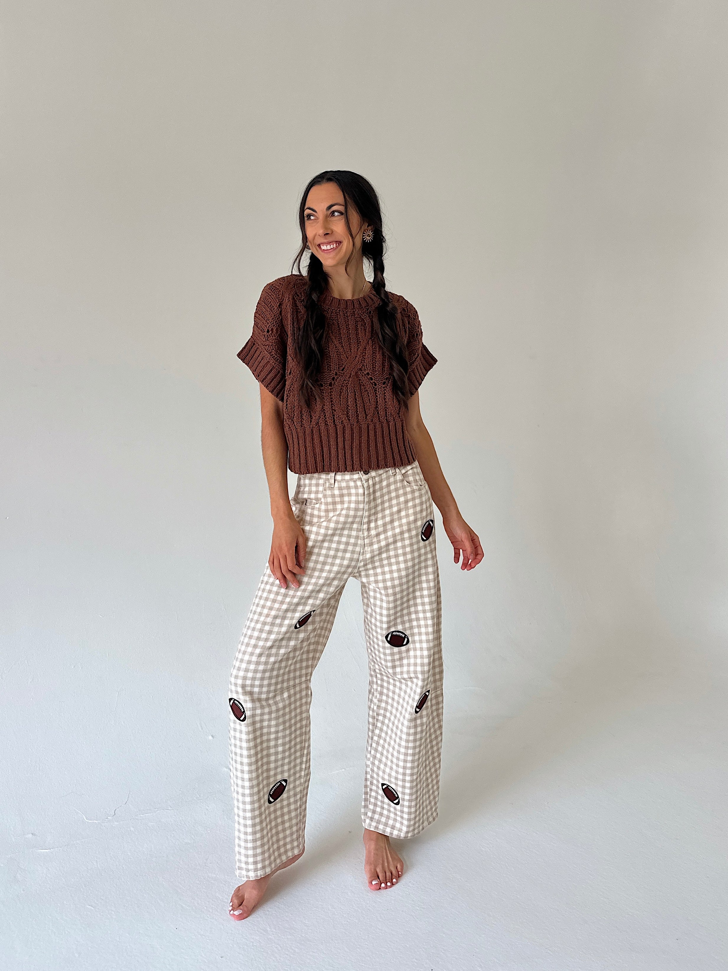 A woman stands barefoot against a plain light gray studio background, wearing a brown short-sleeve knit top and cream gingham wide-leg pants with small dark embroidered motifs.