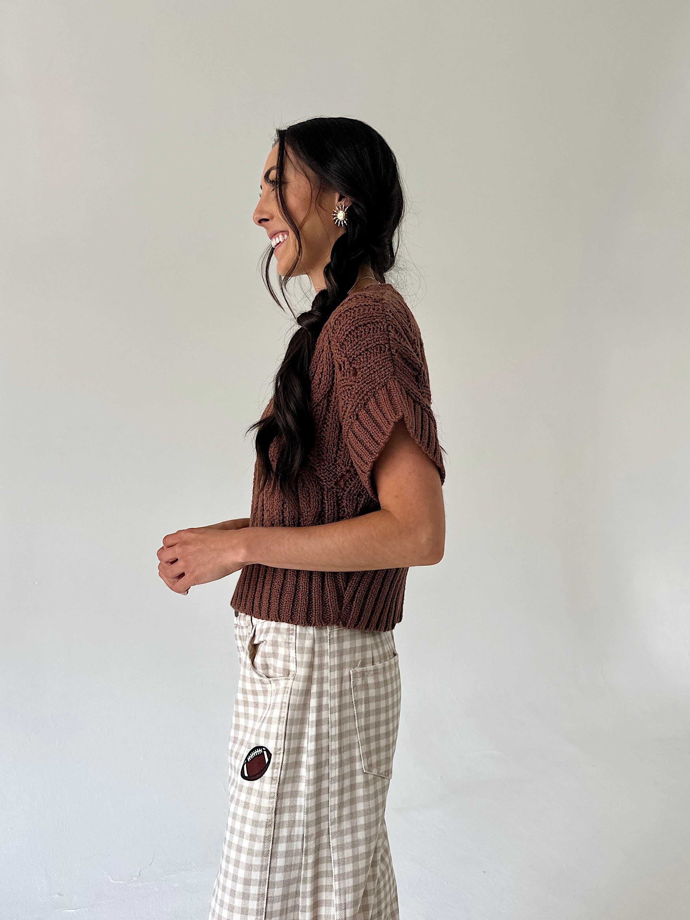 A woman in profile wearing a brown short-sleeve knit top and a beige-and-white gingham pant, standing against a plain light gray wall.