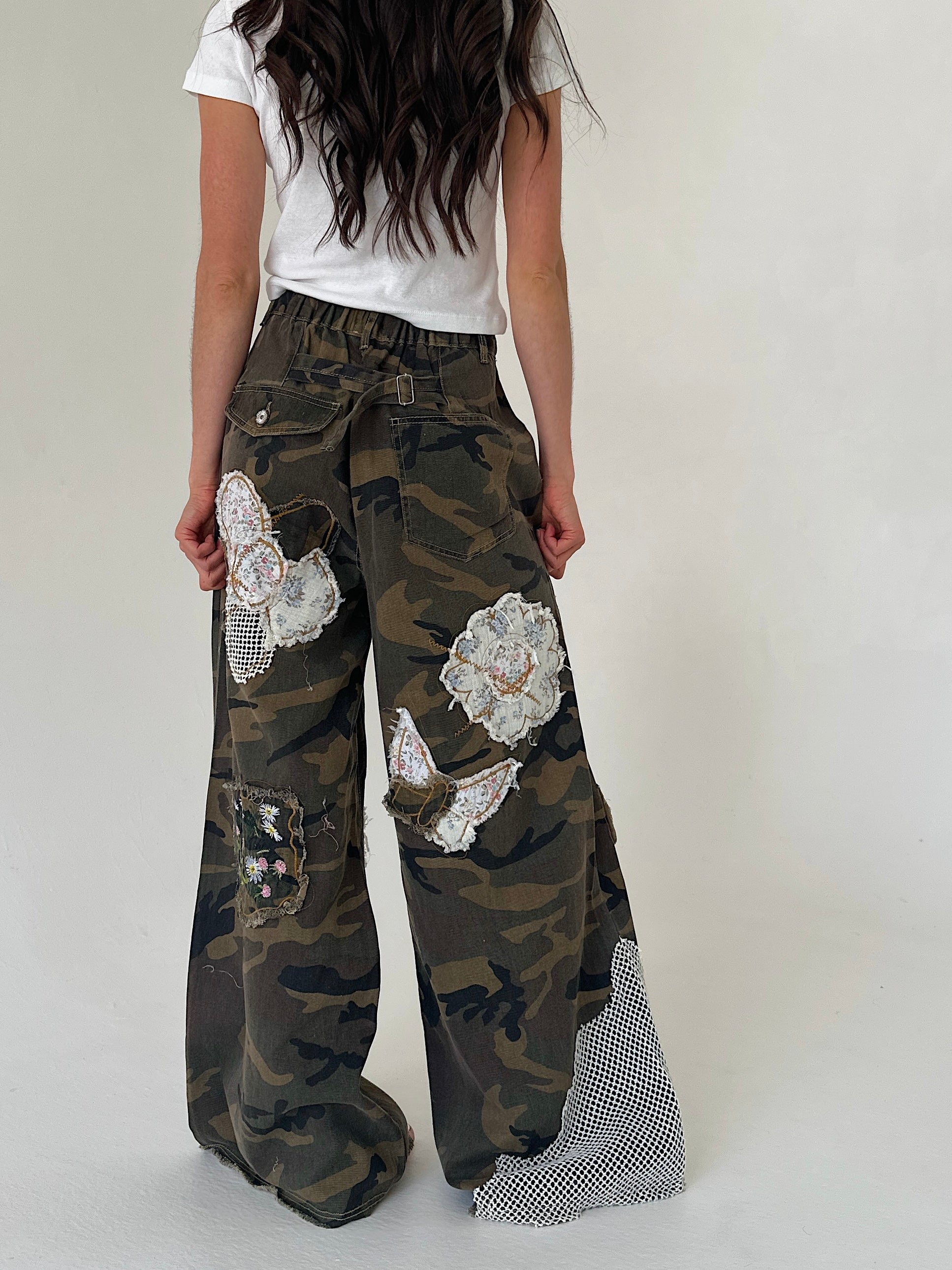 A person shown from the back wearing wide-leg camouflage pants with large white floral applique patches and a mesh fabric panel near the hem, paired with a white T-shirt against a plain light gray studio background.