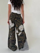 A person shown from the back wearing wide-leg camouflage pants with large white floral applique patches and a mesh fabric panel near the hem, paired with a white T-shirt against a plain light gray studio background.