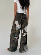 A person shown from the back wearing loose camouflage wide-leg pants with decorative white lace applique patches and a white top, against a plain light gray studio background.