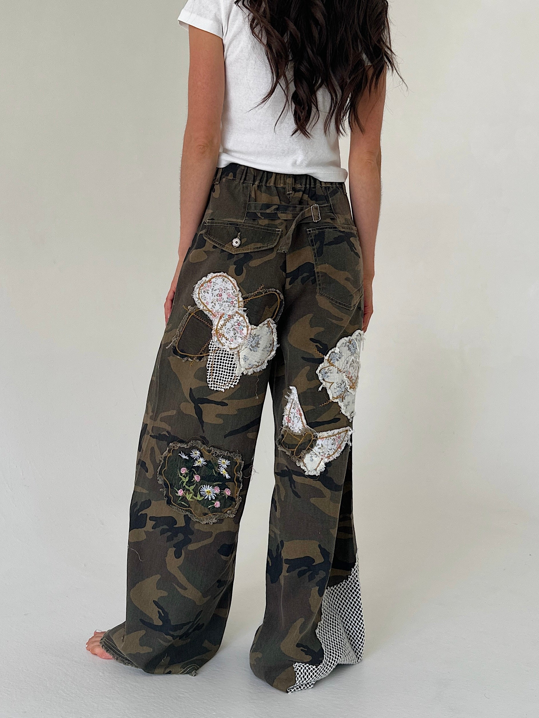 A person shown from the back wearing loose camouflage wide-leg pants with decorative white lace applique patches and a white top, against a plain light gray studio background.
