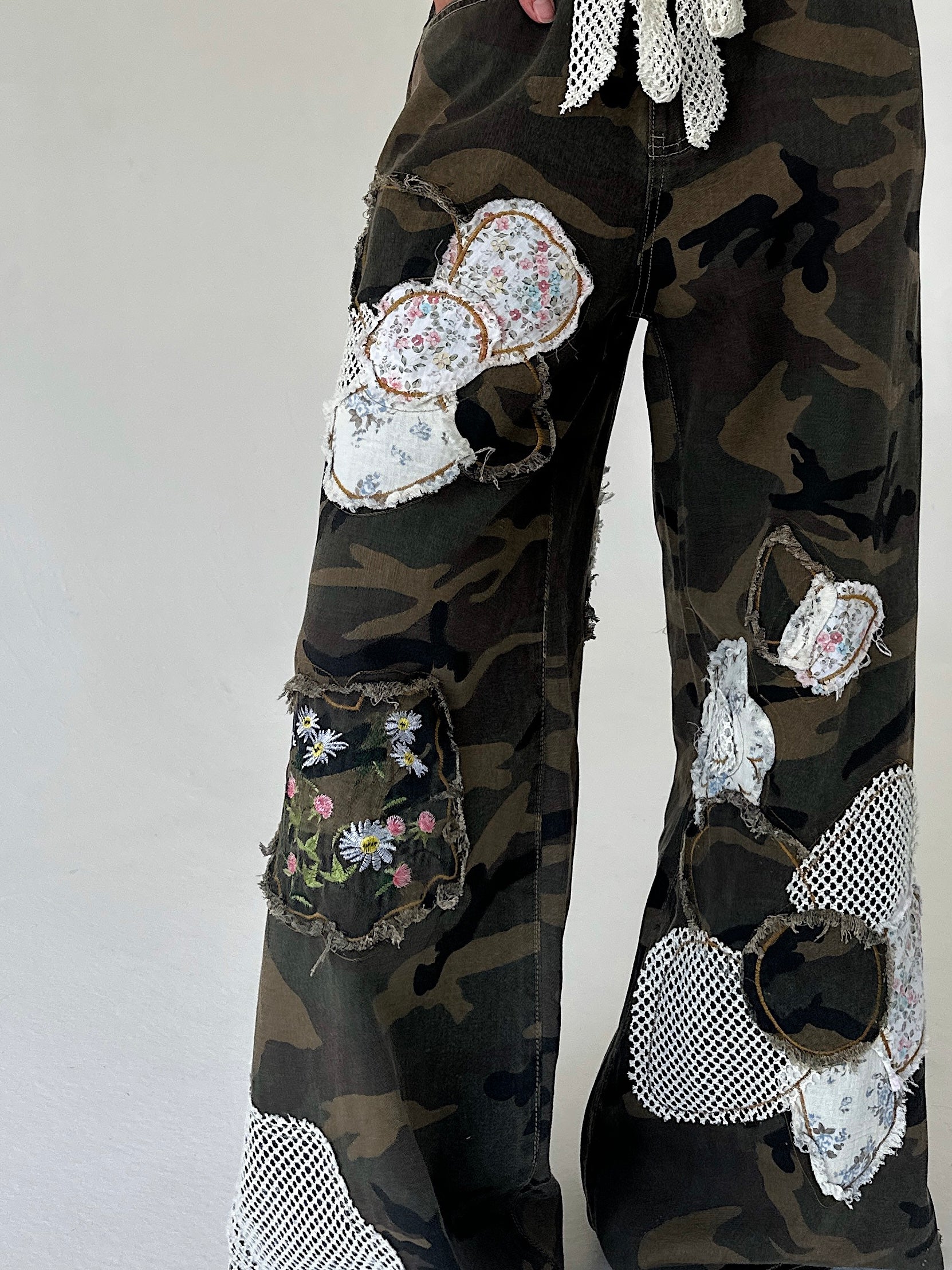 A close-up of a person's lower legs wearing camouflage wide-leg pants heavily embellished with patchwork floral fabric patches, mesh cutouts, and frayed details, set against a plain light gray background.
