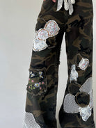 A close-up of a person's lower legs wearing camouflage wide-leg pants heavily embellished with patchwork floral fabric patches, mesh cutouts, and frayed details, set against a plain light gray background.