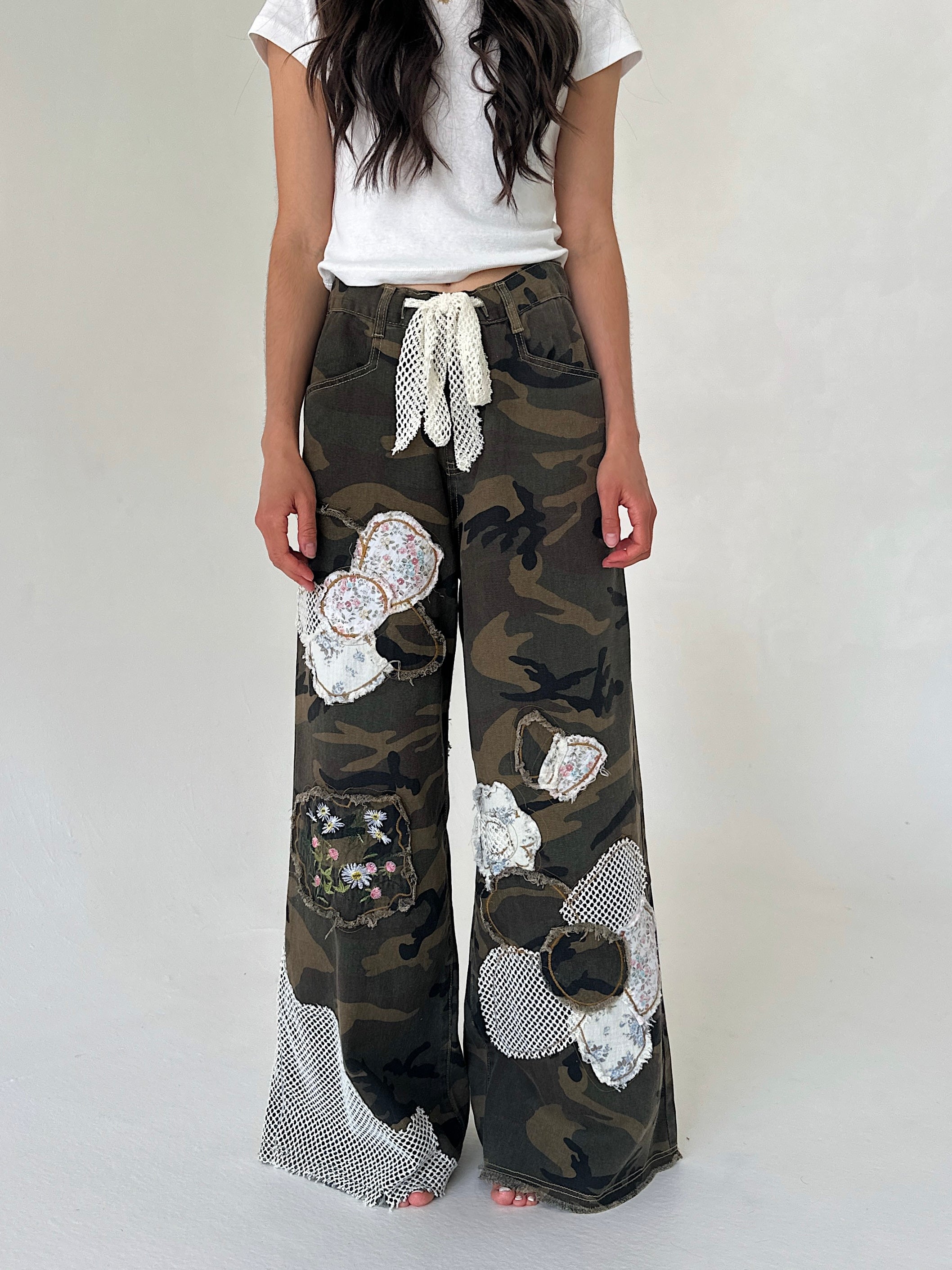 A person wearing loose camouflage wide-leg pants with large white flower appliqués and a white tie waist, paired with a white top, against a plain light background.
