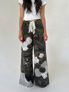 A person wearing loose camouflage wide-leg pants with large white flower appliqués and a white tie waist, paired with a white top, against a plain light background.