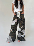 A person wearing loose camouflage wide-leg pants with large white sequined flower appliqués, a white drawstring waist, and mesh hem detailing, paired with a white cropped T-shirt against a plain light gray studio background.