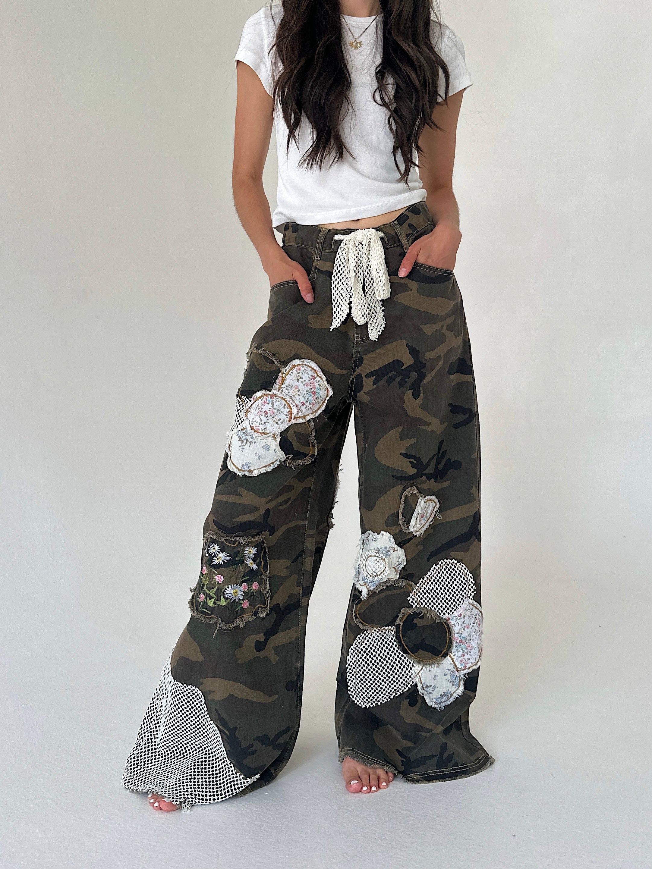 A person wearing loose camouflage wide-leg pants with large white sequined flower appliqués, a white drawstring waist, and mesh hem detailing, paired with a white cropped T-shirt against a plain light gray studio background.