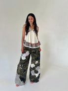 A woman modeling a sleeveless cream top with a ruffled hem and wide-leg camouflage pants featuring white mesh patchwork details, standing against a plain light gray studio background.
