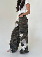 A model wearing a pair of wide-leg camouflage cargo pants decorated with floral patches and mesh trim, paired with a white t-shirt against a plain light gray background.