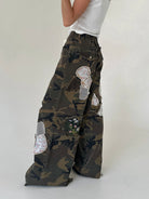 A person wearing wide-leg camouflage cargo pants with large decorative floral patches, paired with a white sleeveless top, photographed against a plain light gray studio background.