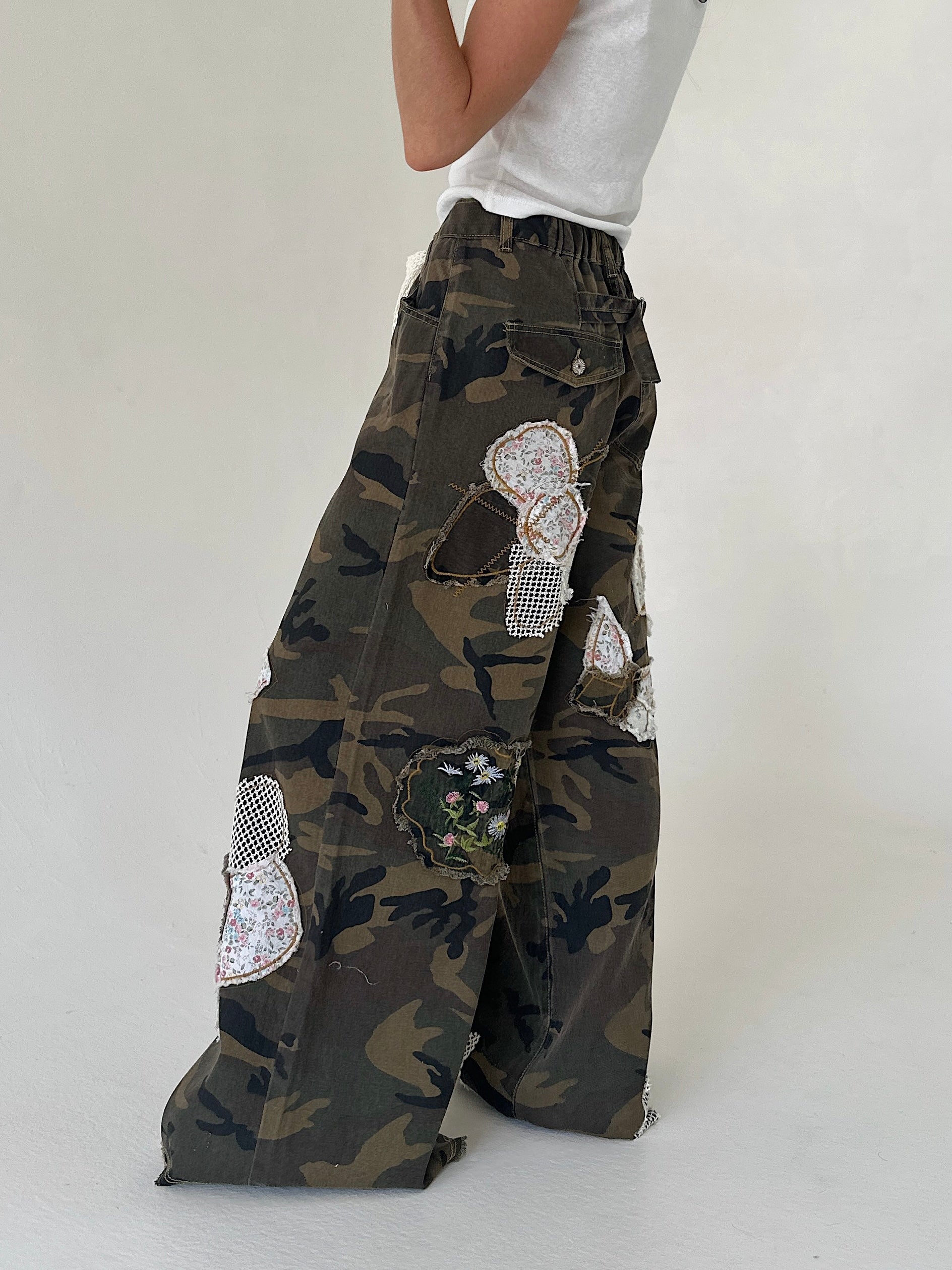 A person wearing wide-leg camouflage cargo pants with large decorative floral patches, paired with a white sleeveless top, photographed against a plain light gray studio background.