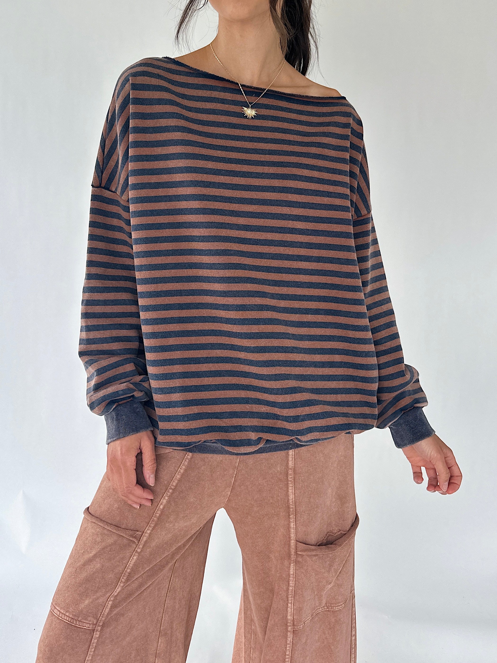 A woman wearing a loose, off-the-shoulder long-sleeve top with horizontal dark brown and navy stripes, paired with dusty rose wide-leg pants, against a plain light background.