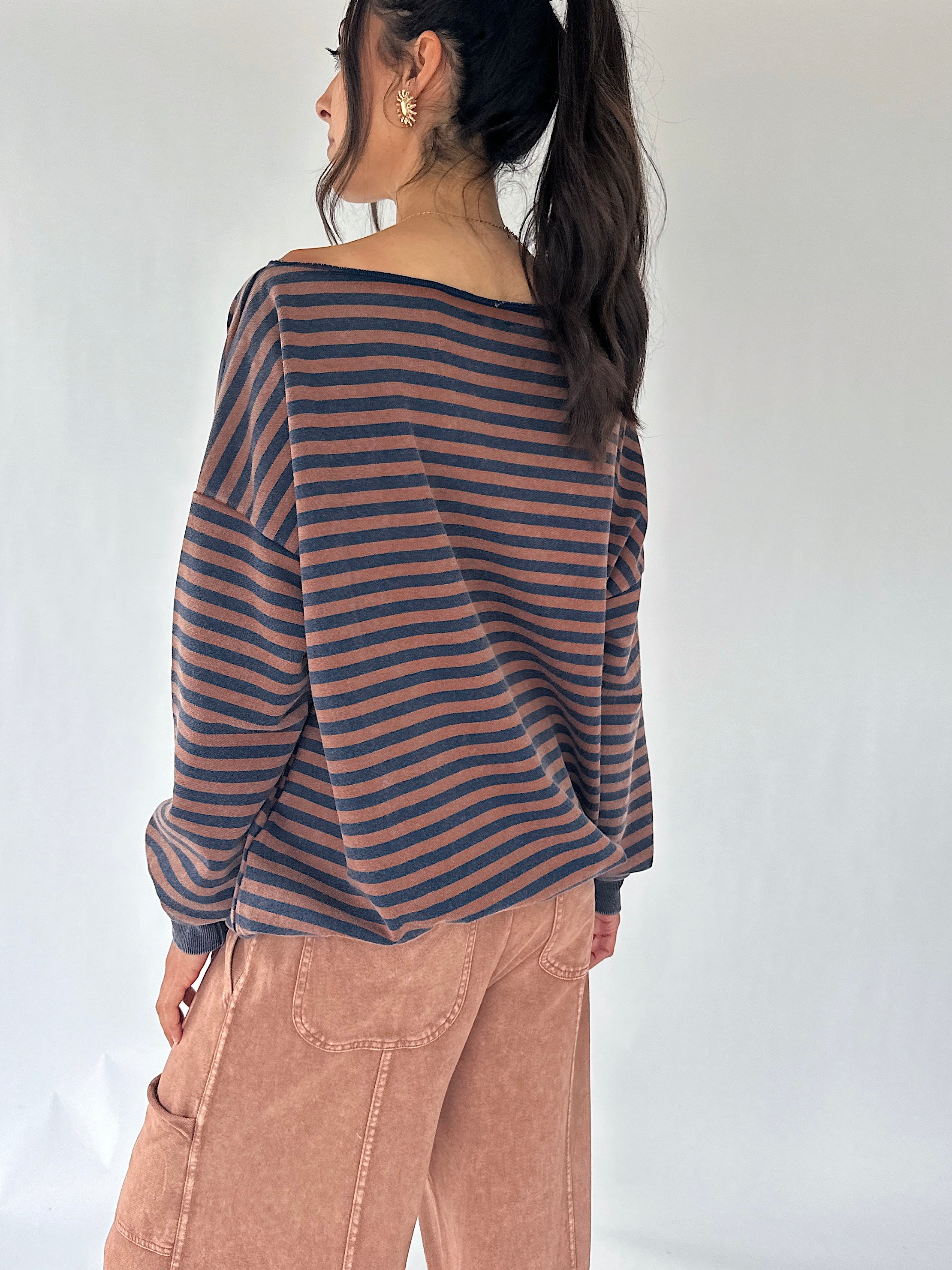 A woman shown from the back wearing a loose, off-the-shoulder long-sleeve top with horizontal dark brown and dusty navy stripes, paired with dusty brown wide leg pants, against a plain white studio background.