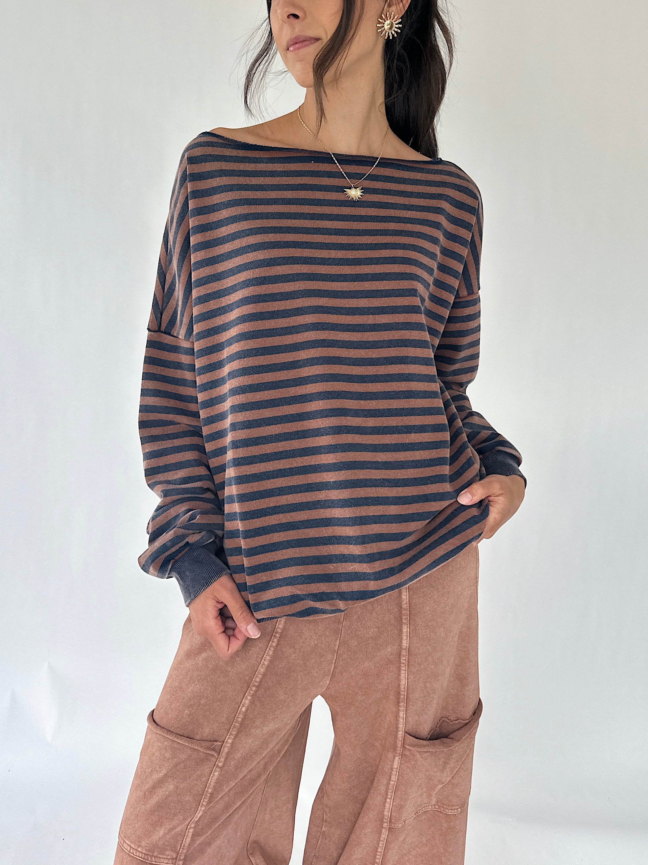 A woman wearing a loose, off-the-shoulder long-sleeve top with horizontal dark brown and muted blue stripes, paired with brown wide-leg pants, against a plain white studio background.