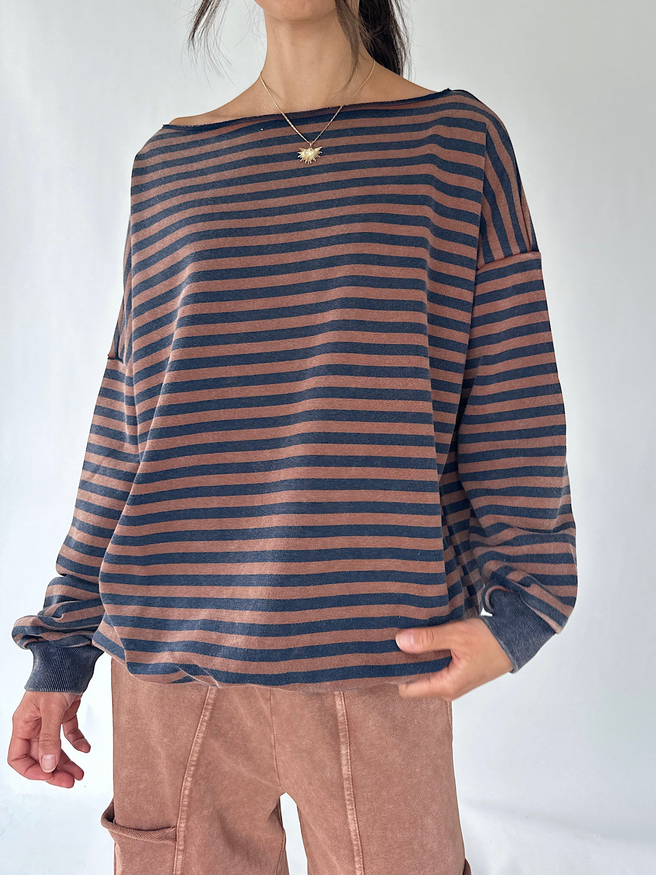 A woman wearing a loose, long-sleeve striped top with horizontal navy and dusty rose-brown stripes, paired with brown pants, against a plain light background.