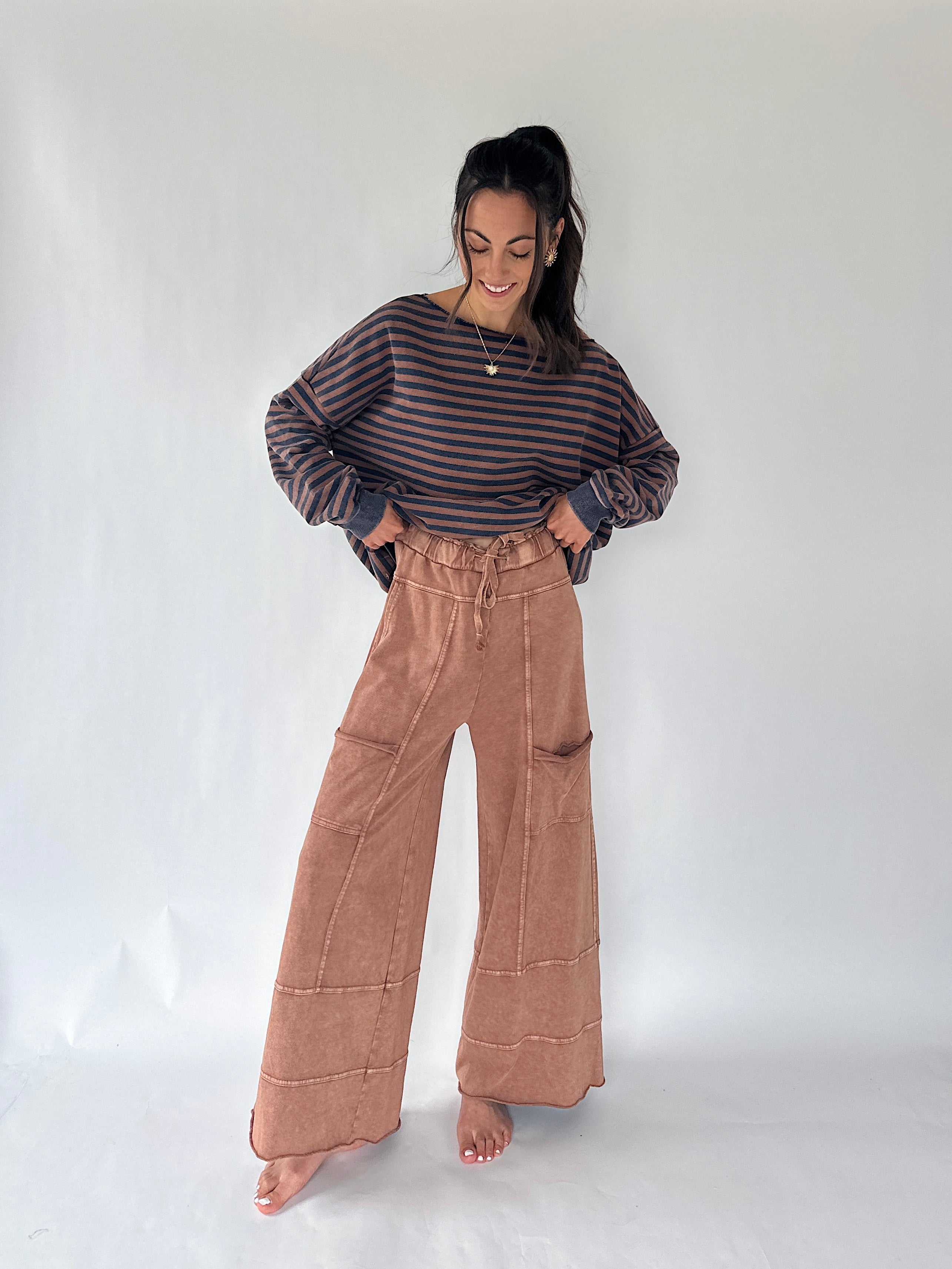 A woman stands barefoot against a plain light gray studio background, wearing a dark striped long-sleeve top and high-waisted wide-leg tan pants.
