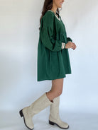 Person wearing a green dress and white knee-high boots against a white background side view