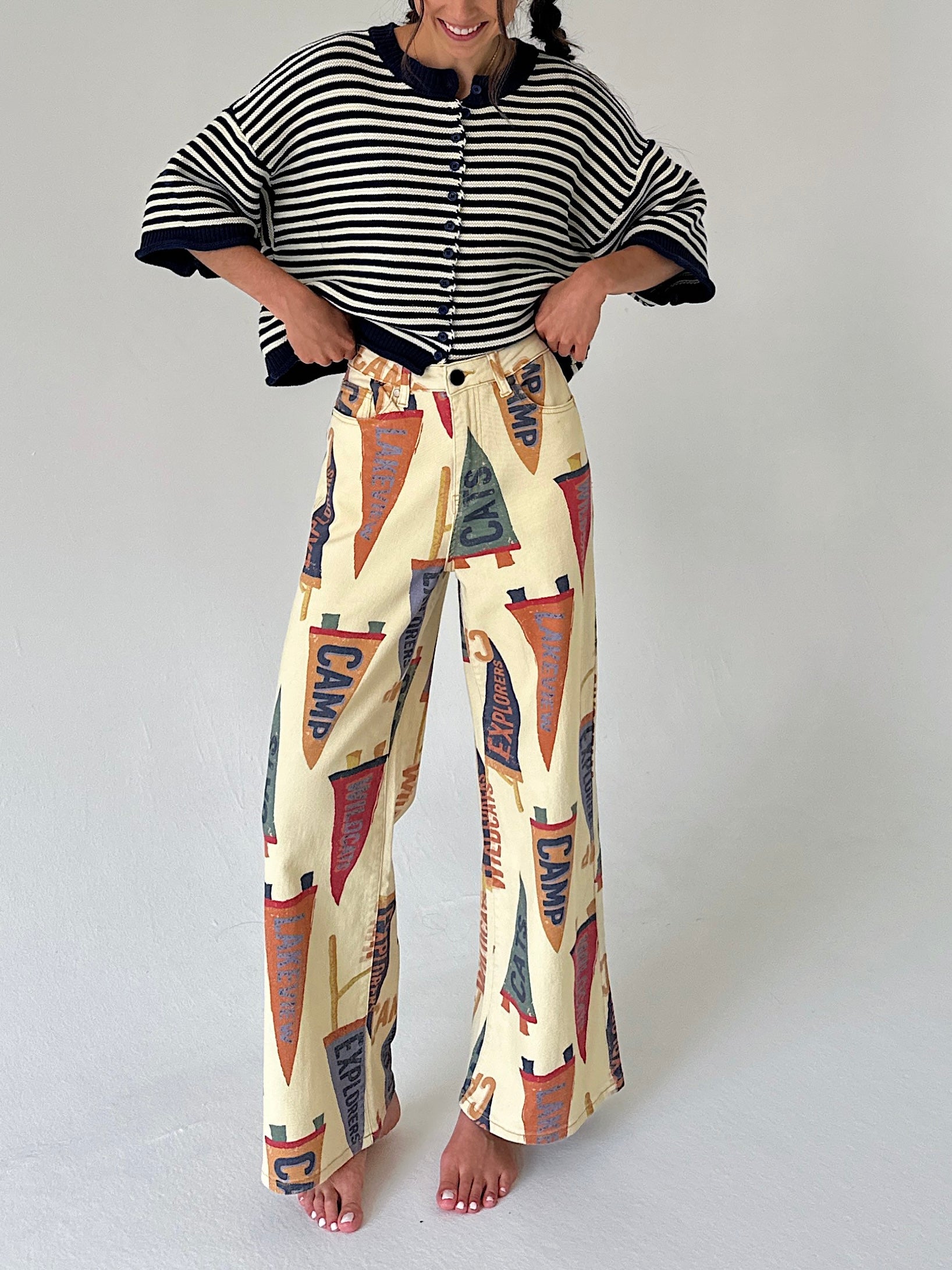 A woman modeling a two-piece outfit consisting of a blue and white striped short-sleeve cardigan and high-waisted, wide-leg pants featuring a vintage pennant flag print on a cream background.
