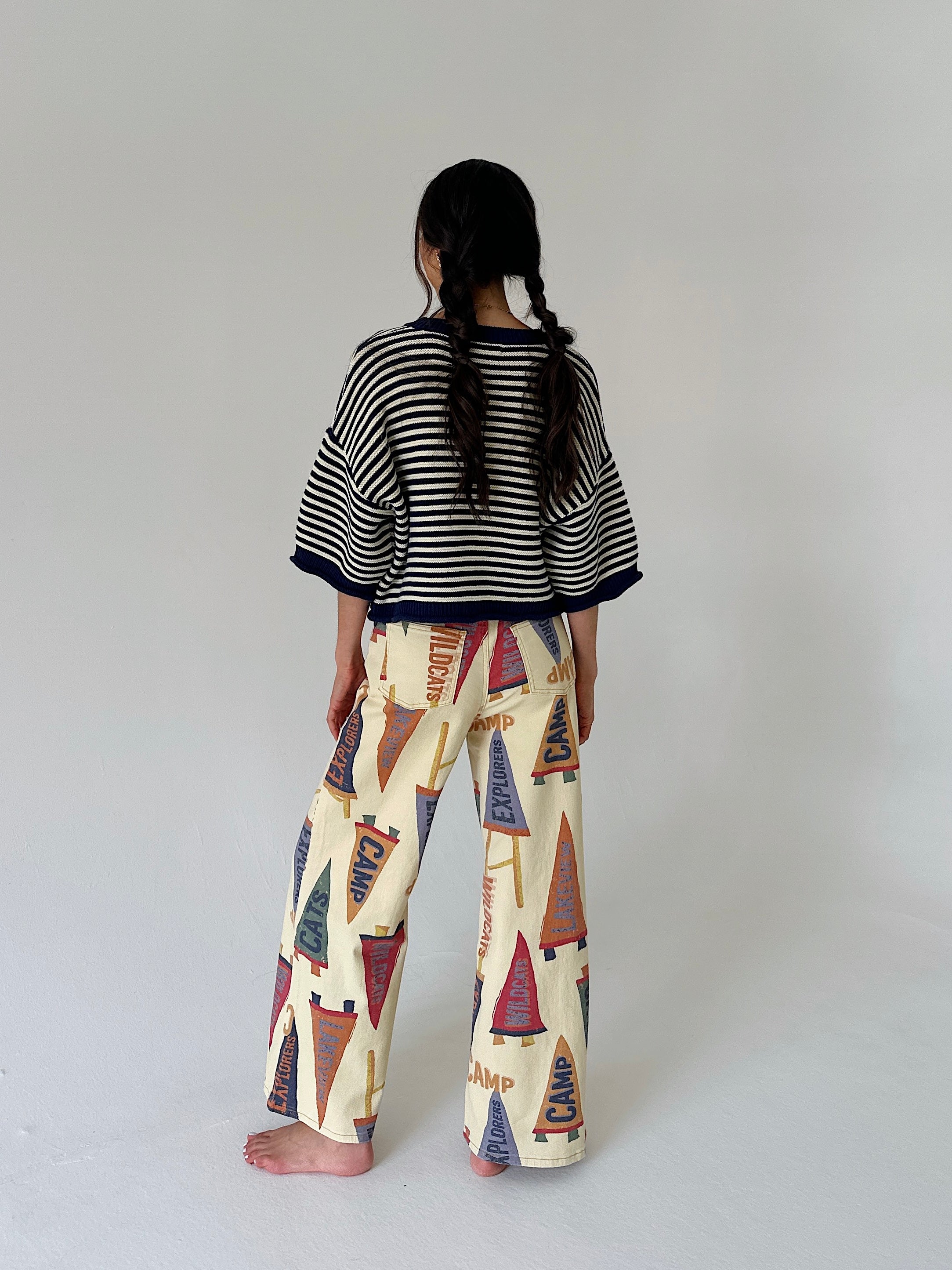 A child model shown from behind wearing a black and white striped short-sleeve top and cream wide-leg pants with a colorful pennant flag pattern, standing against a plain light gray studio background.