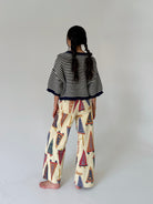 A child model shown from behind wearing a black and white striped short-sleeve top and cream wide-leg pants with a colorful pennant flag pattern, standing against a plain light gray studio background.