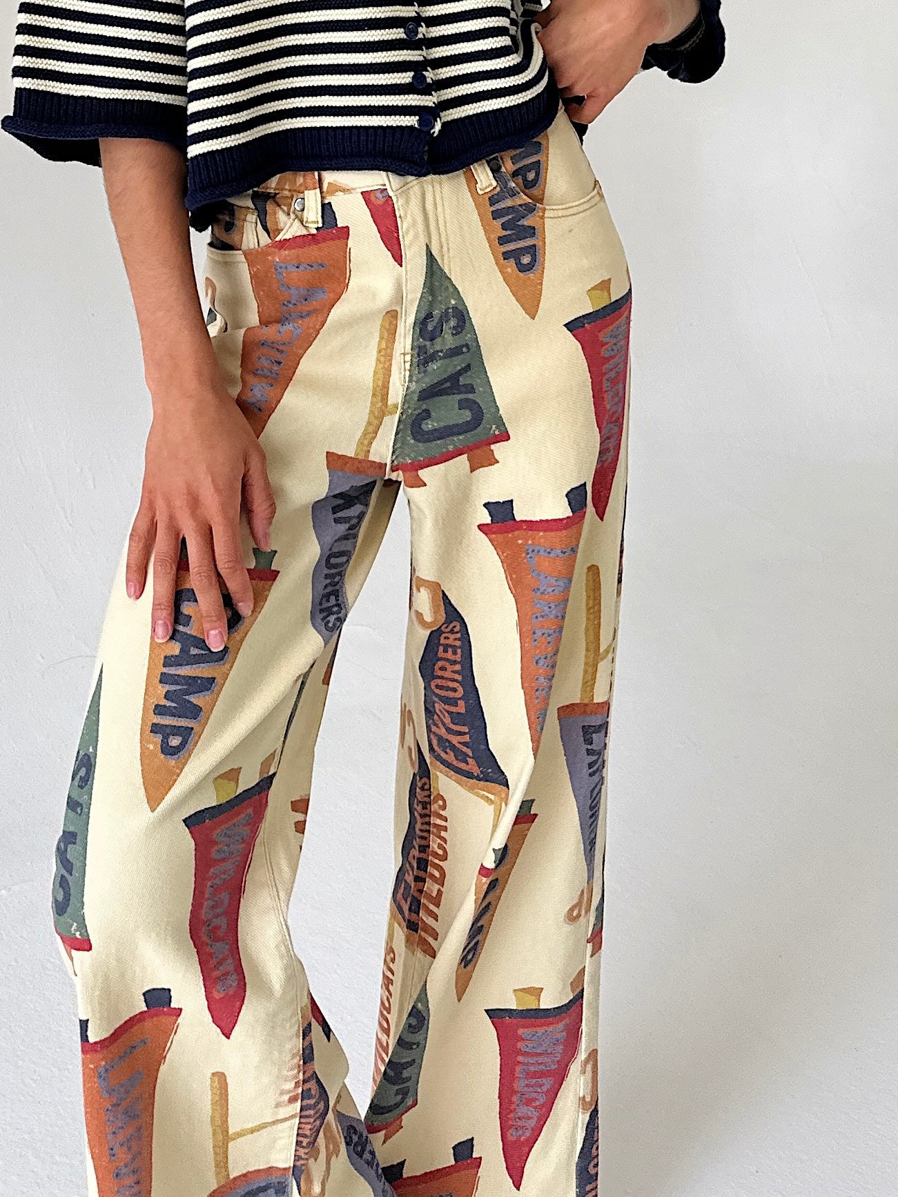 A person wearing wide-leg cream pants with an allover vintage pennant flag print in red, blue, orange, and brown, paired with a black-and-white striped top against a plain light background.