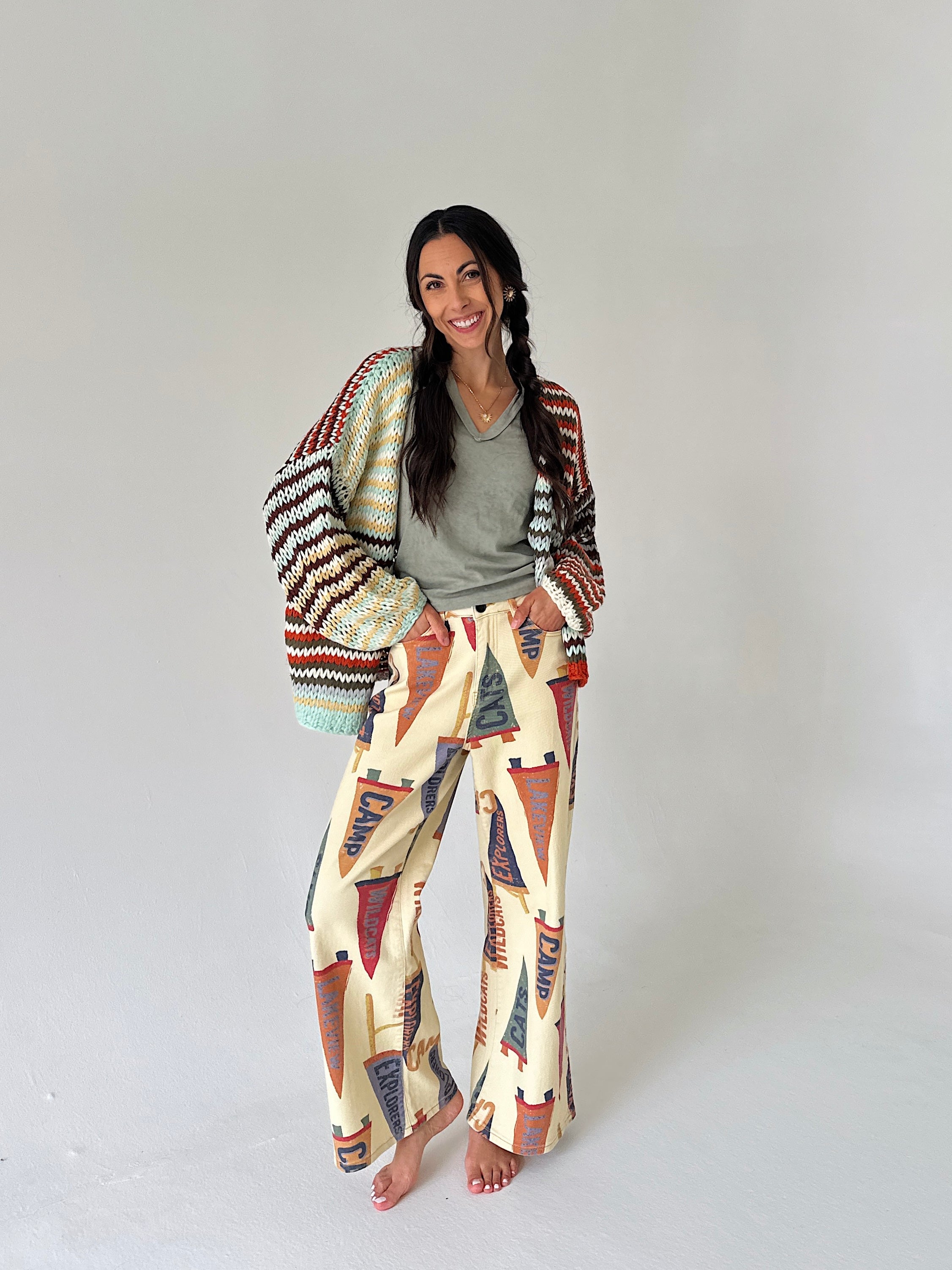 A woman stands barefoot against a plain light gray studio background, wearing cream wide-leg pants with a colorful pennant print, a green top, and a multicolor knit cardigan.