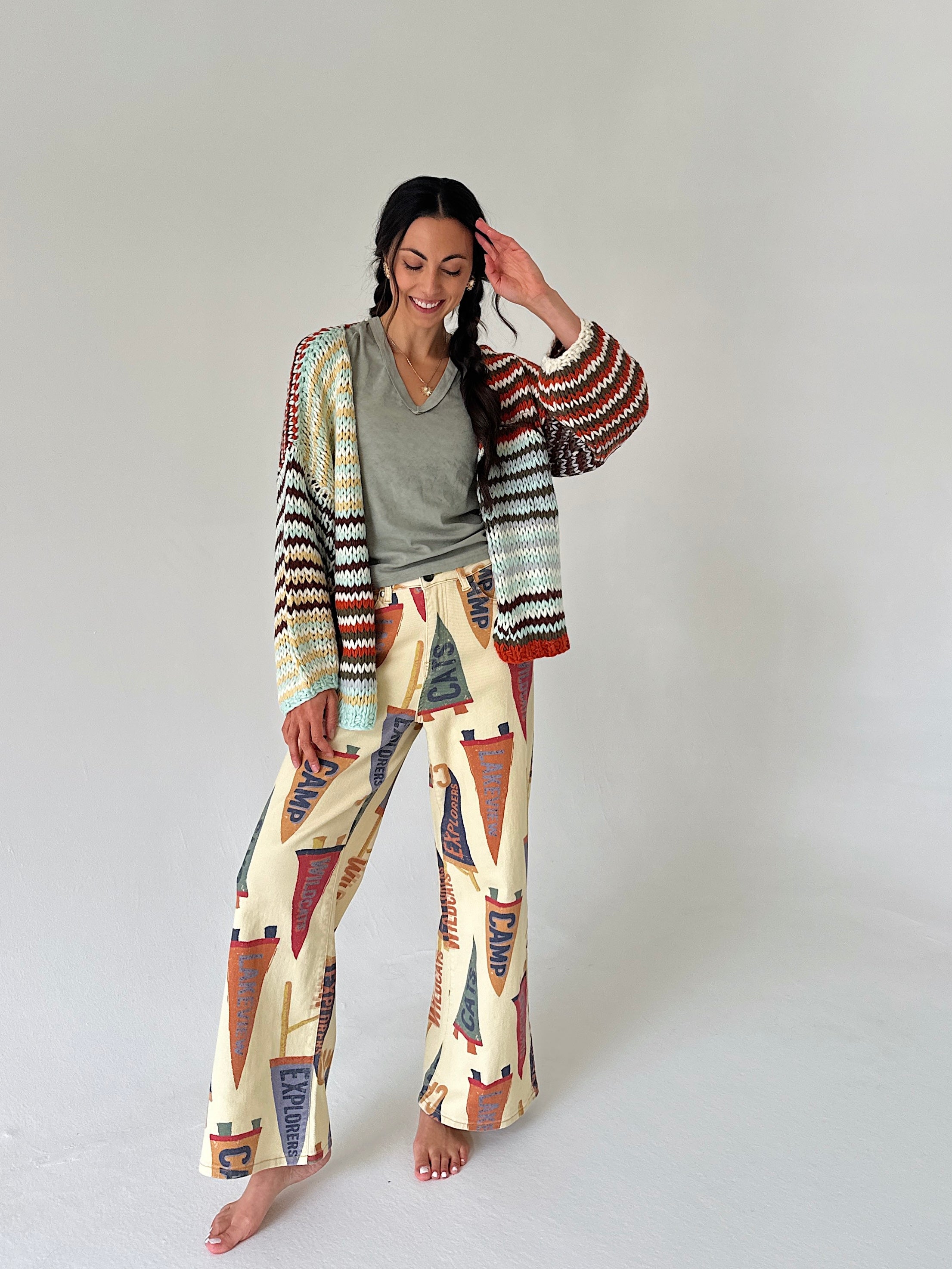 A woman stands barefoot against a plain light gray studio background, wearing a colorful striped knit cardigan and wide-leg pants set with a cream base and pennant multicolor patterns in rust, navy, olive, and black.