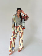 A woman stands barefoot against a plain light gray studio background, wearing a colorful striped knit cardigan and wide-leg pants set with a cream base and pennant multicolor patterns in rust, navy, olive, and black.