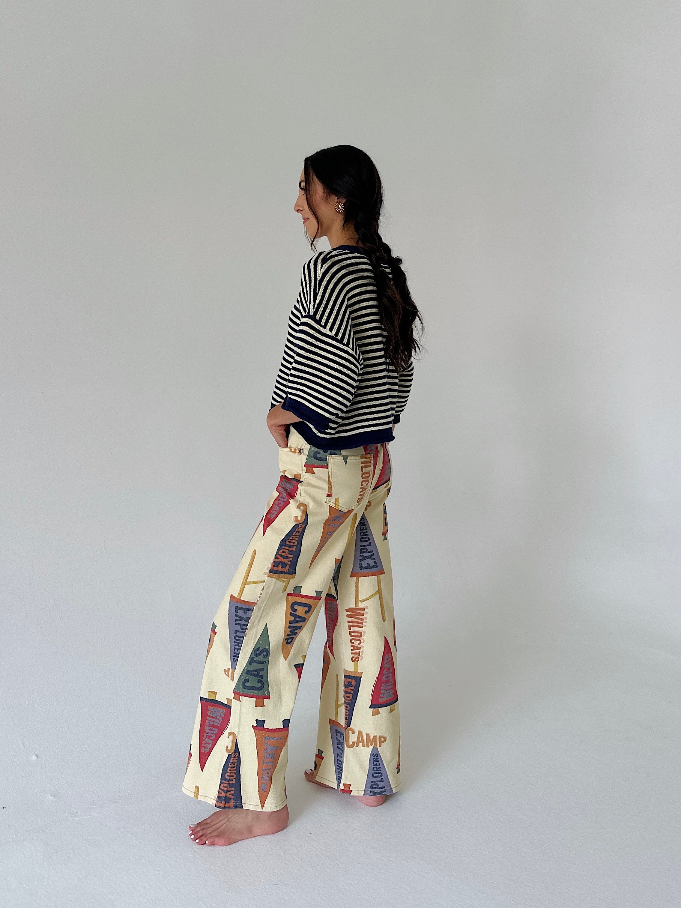 A woman stands barefoot in profile wearing a black and white striped cardigan and cream wide-leg pants with a colorful vintage flag print, set against a plain light gray studio background.