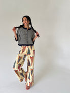 A woman modeling a two-piece outfit consisting of a navy and white striped short-sleeve cardigan with a button-up front and cream wide-leg pants featuring a colorful pennant flag pattern, set against a plain light gray studio background.