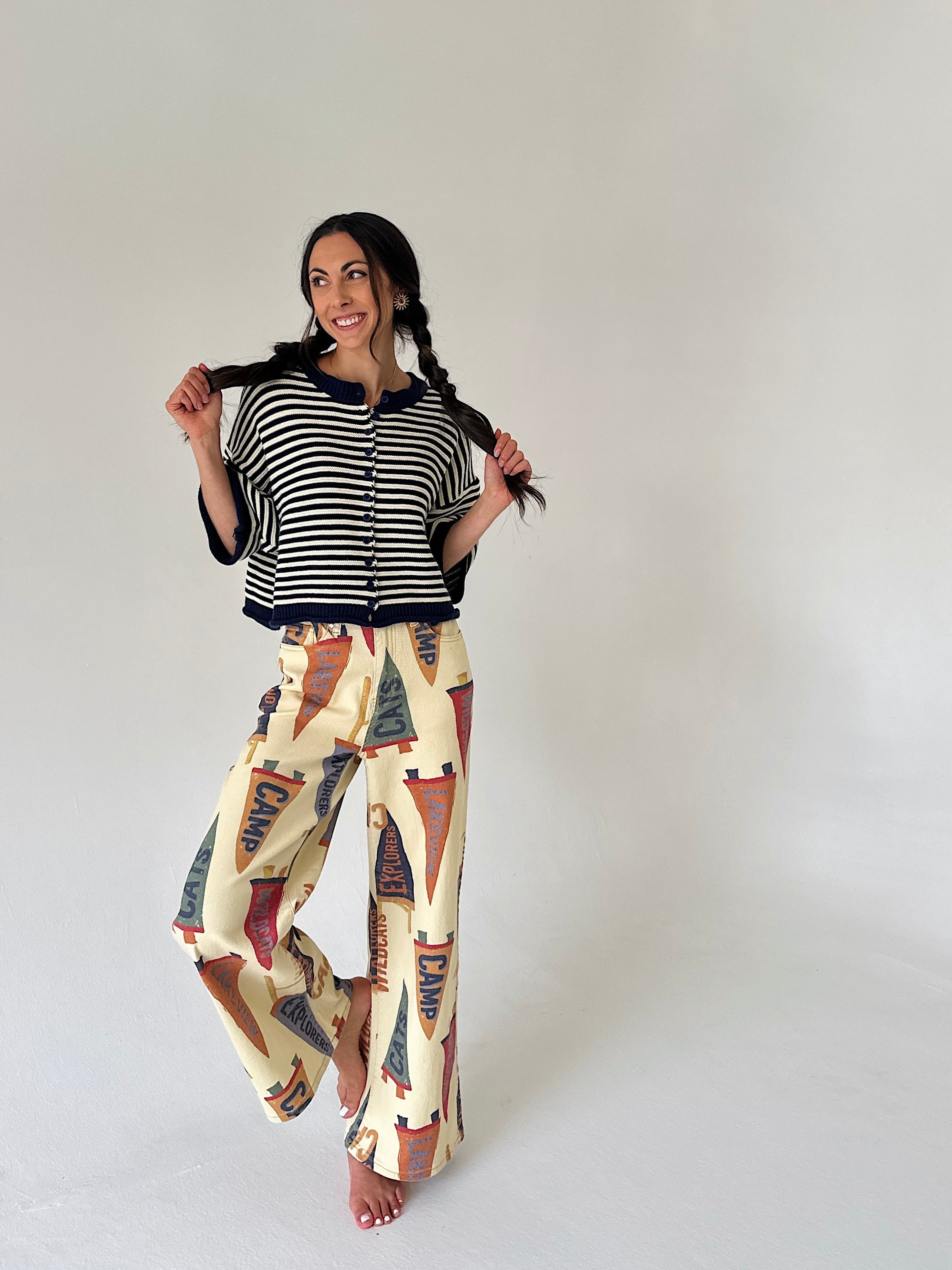 A woman modeling a two-piece outfit consisting of a navy and white striped short-sleeve cardigan with a button-up front and cream wide-leg pants featuring a colorful pennant flag pattern, set against a plain light gray studio background.