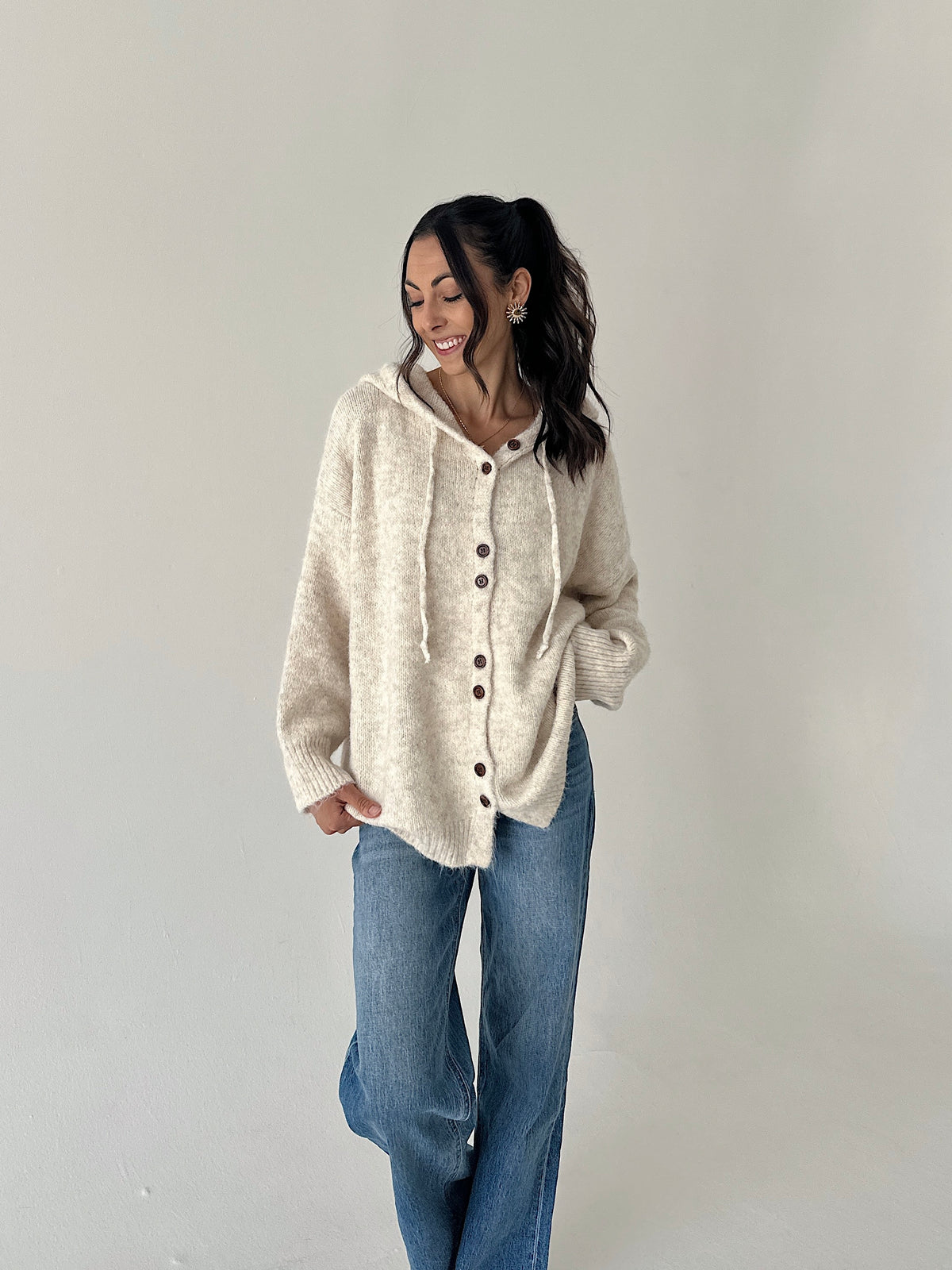 A woman wearing a cream-colored, fuzzy button-up hooded sweater and blue jeans, standing against a plain light gray studio background.