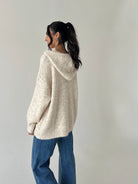 A woman wearing a cream-colored, oversized knit hooded sweater and blue jeans, shown from a side-back angle against a plain light gray wall.