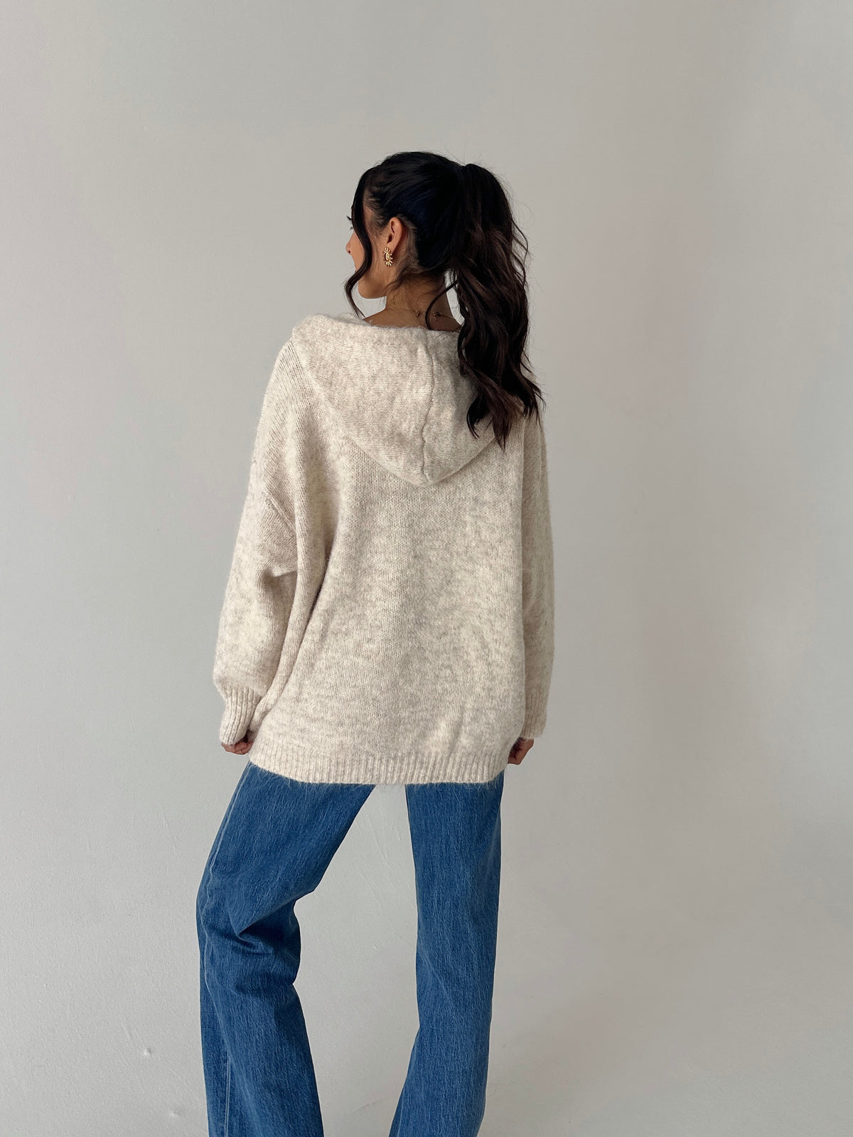 A woman shown from the back wearing a cream-colored, oversized knit hoodie sweater and blue jeans, standing against a plain light gray wall.
