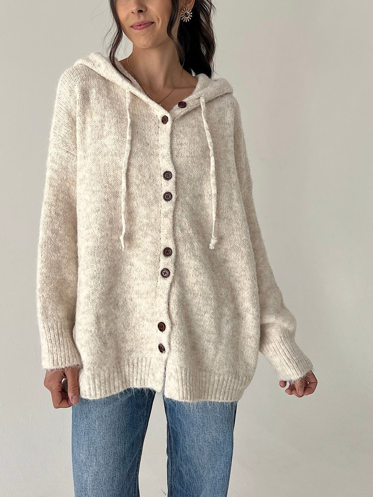 A woman wearing a cream-colored, fuzzy knit hooded button-up sweater with brown buttons and matching drawstrings, paired with blue jeans, against a plain light gray background.