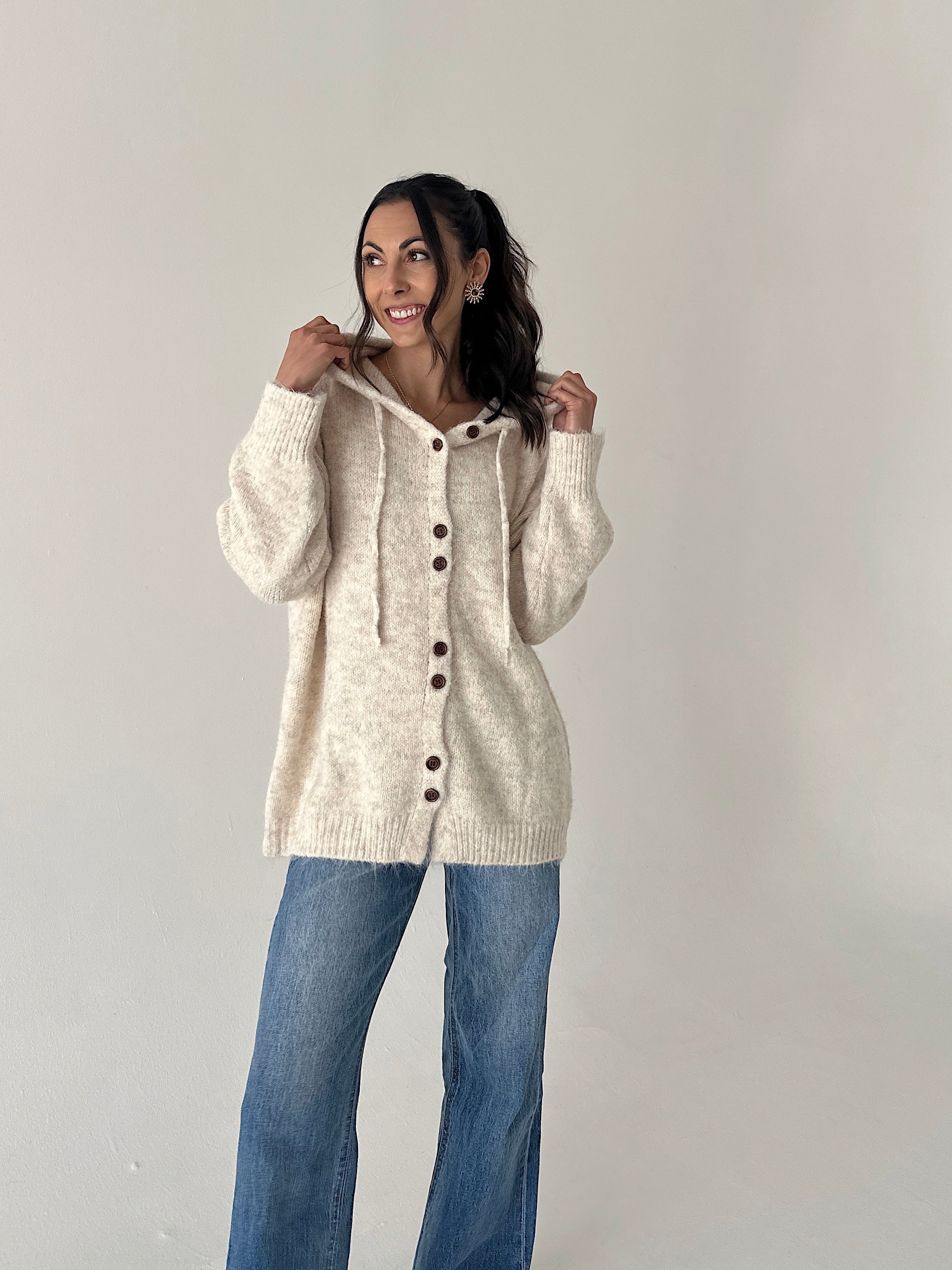 A woman modeling a cream-colored, oversized knit cardigan with a hood and front buttons, paired with blue jeans, against a plain light gray studio background.