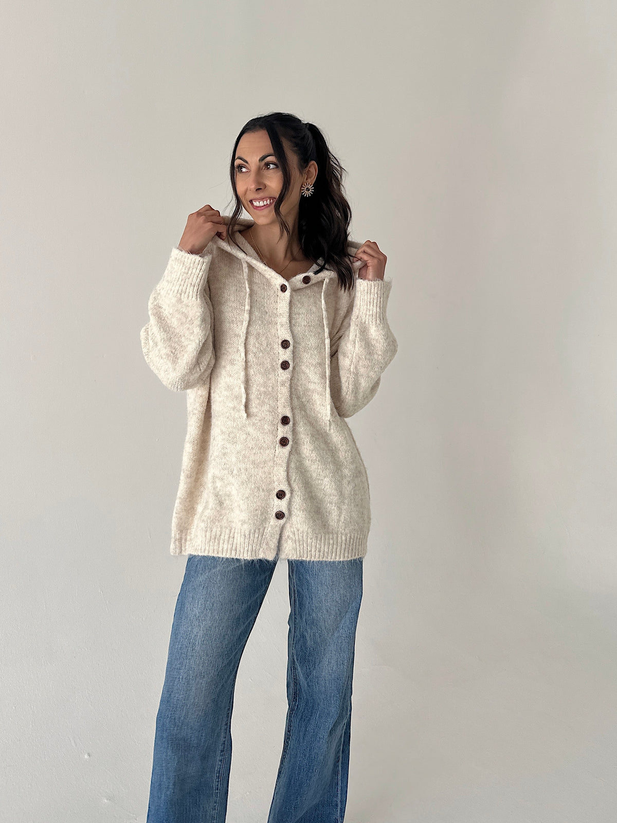 A woman modeling a cream-colored, oversized knit cardigan with a hood and front buttons, paired with blue jeans, against a plain light gray studio background.