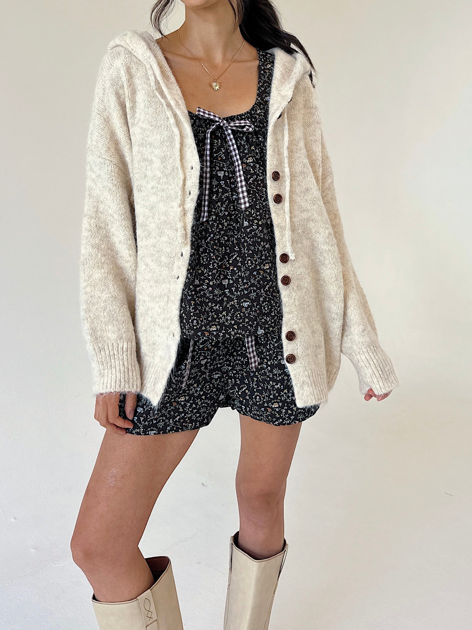 A model wearing a cream-colored, fuzzy knit cardigan over a floral tank and short set, paired with cream-colored knee-high boots, against a plain off-white background.